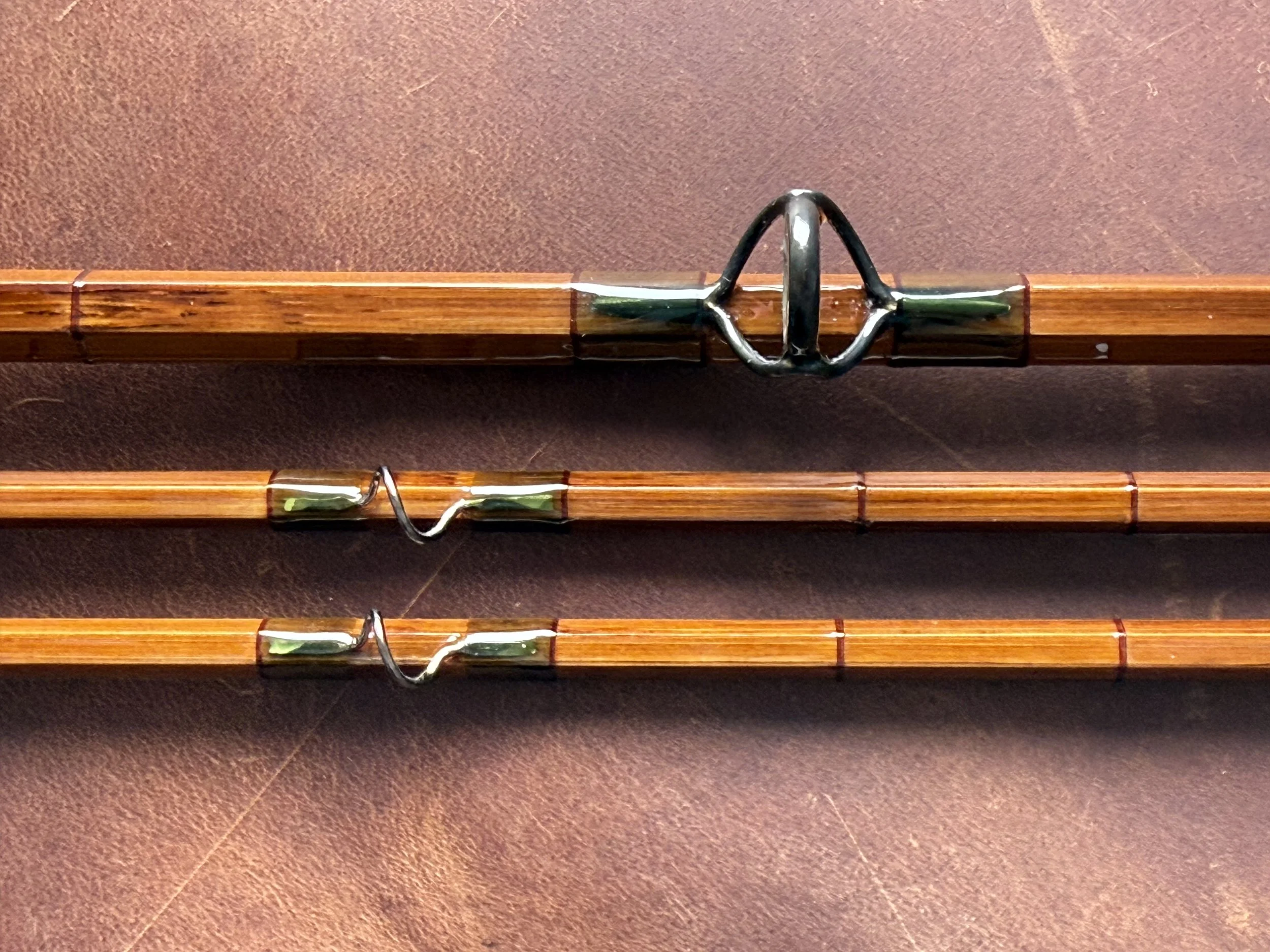 Close-up of three bamboo fly rod sections displaying a blued stripping guide on the butt and two smaller snake guides on the twin tip sections, with translucent olive green and copper thread wrappings.