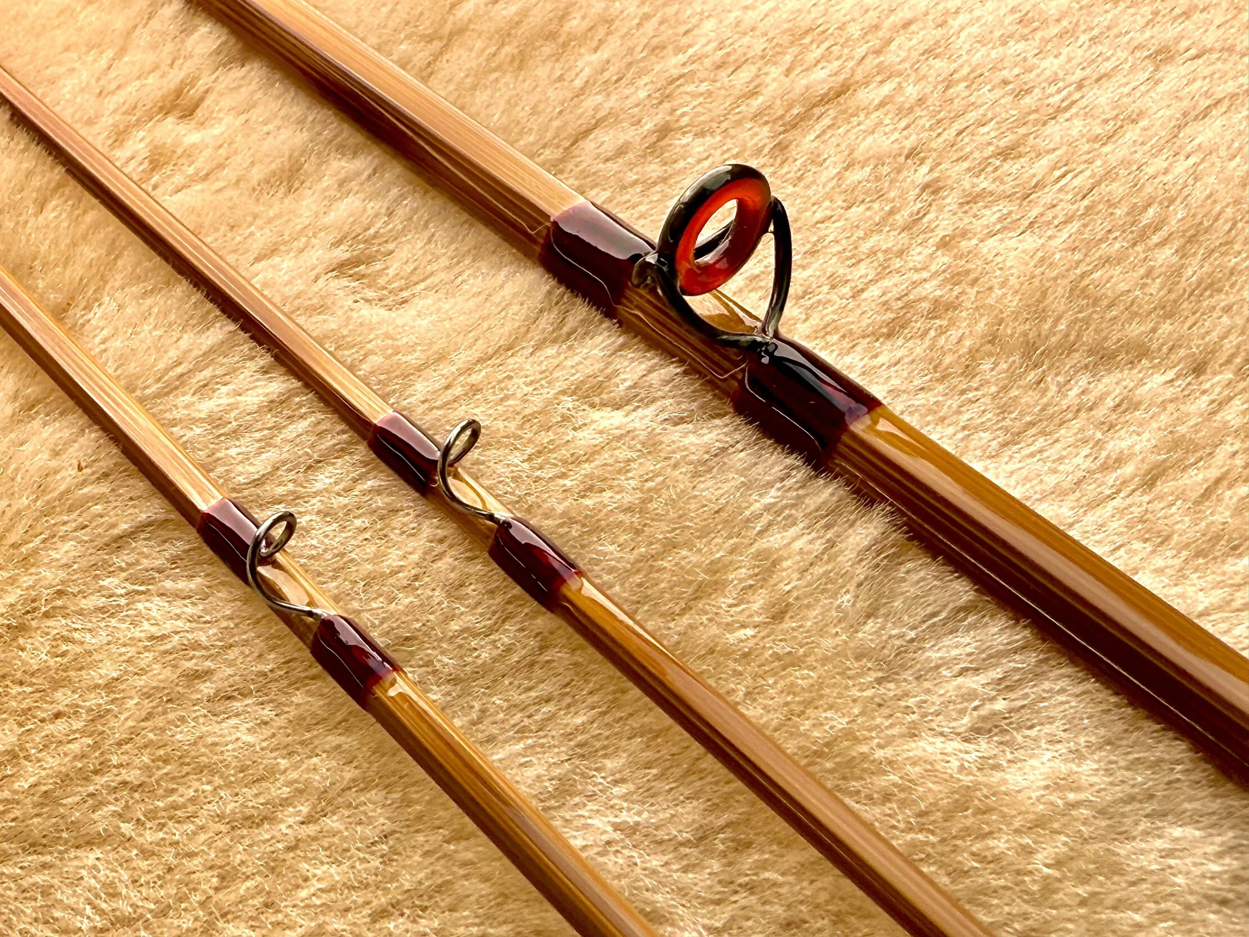 Three bamboo fly rod sections laid diagonally on a warm golden sheepskin surface, showing flamed blanks with a stripping guide fitted with a red agate ring insert and burgundy thread wraps, plus two smaller snake guides with matching burgundy thread 