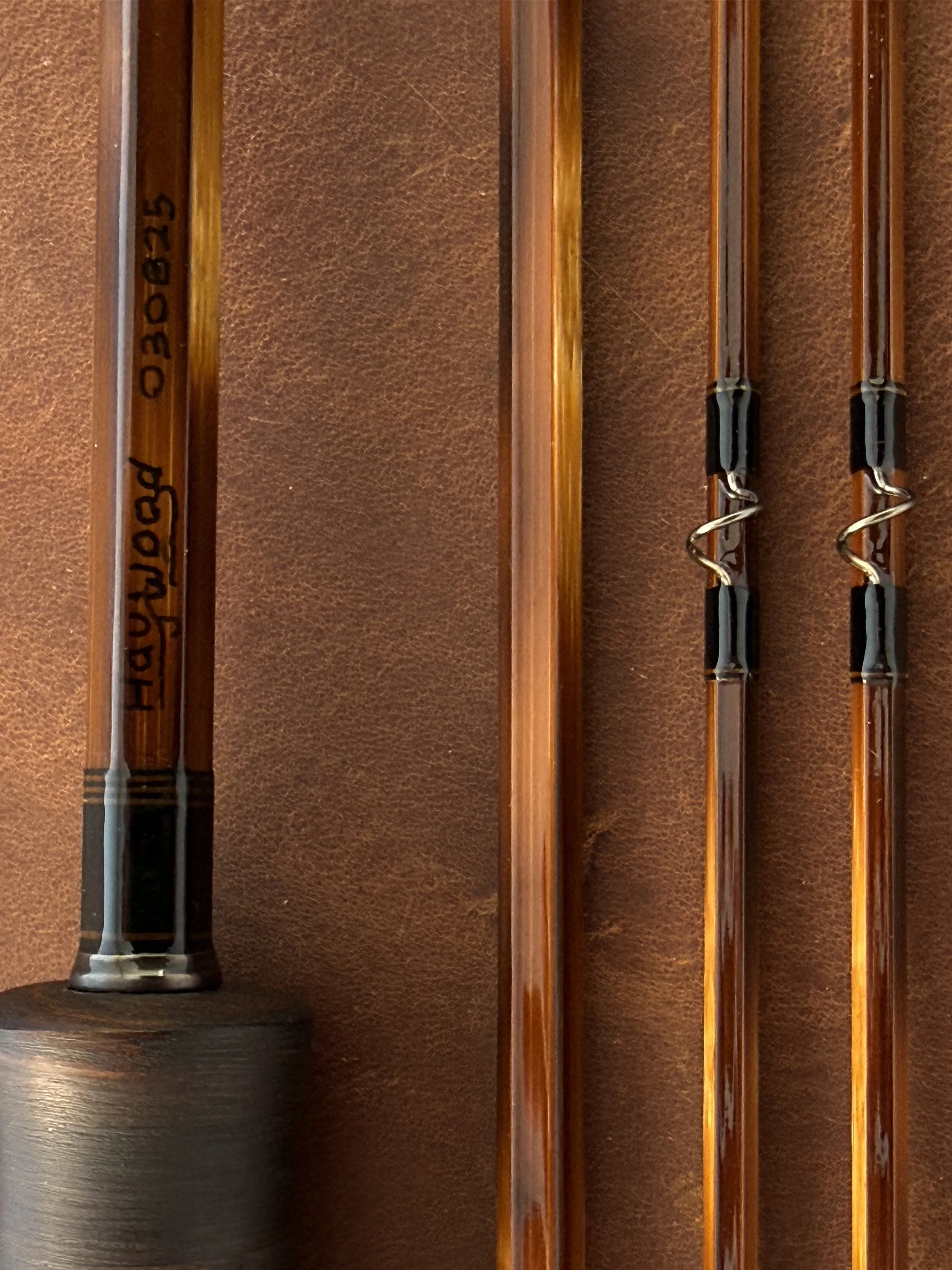 Close-up of a Haywood fly rod butt section marked "Haywood 030825" alongside three heavily flamed rod sections with dark nickel snake guides with black thread wrappings, displayed vertically against a brown leather background.