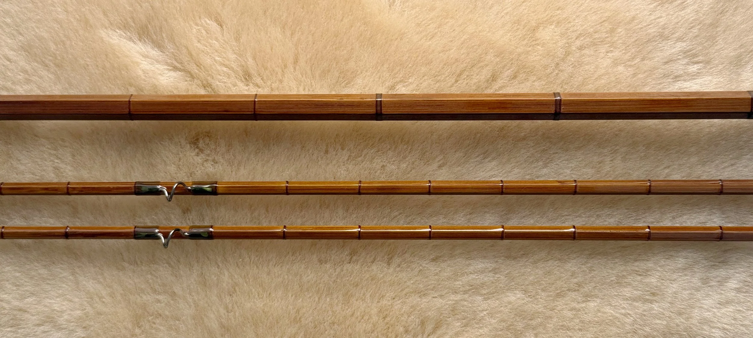 Three sections of a handcrafted bamboo fly rod laid flat on a light fur or sheepskin surface, displaying the hexagonal cane construction, evenly spaced nodes, olive green and copper wrappings with intermediate and signature wraps and snake guides alo