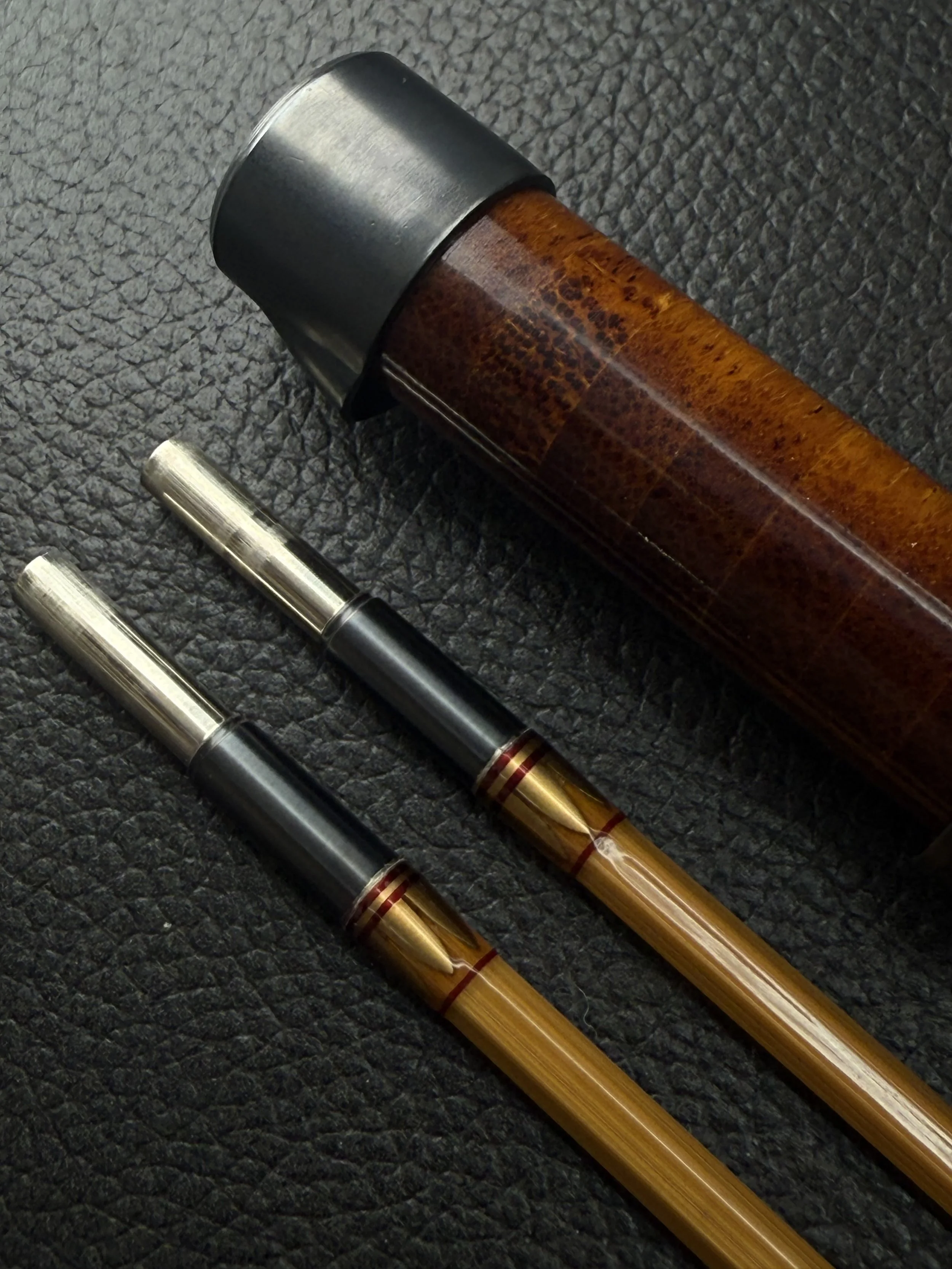 Close-up of a handcrafted bamboo fly rod reel seat featuring a flamed bamboo insert with a blued nickel silver end cap, beside two blonde bamboo rod sections with blued ferrule sleeves, antique gold main wraps and red thread accent wraps, on a dark l