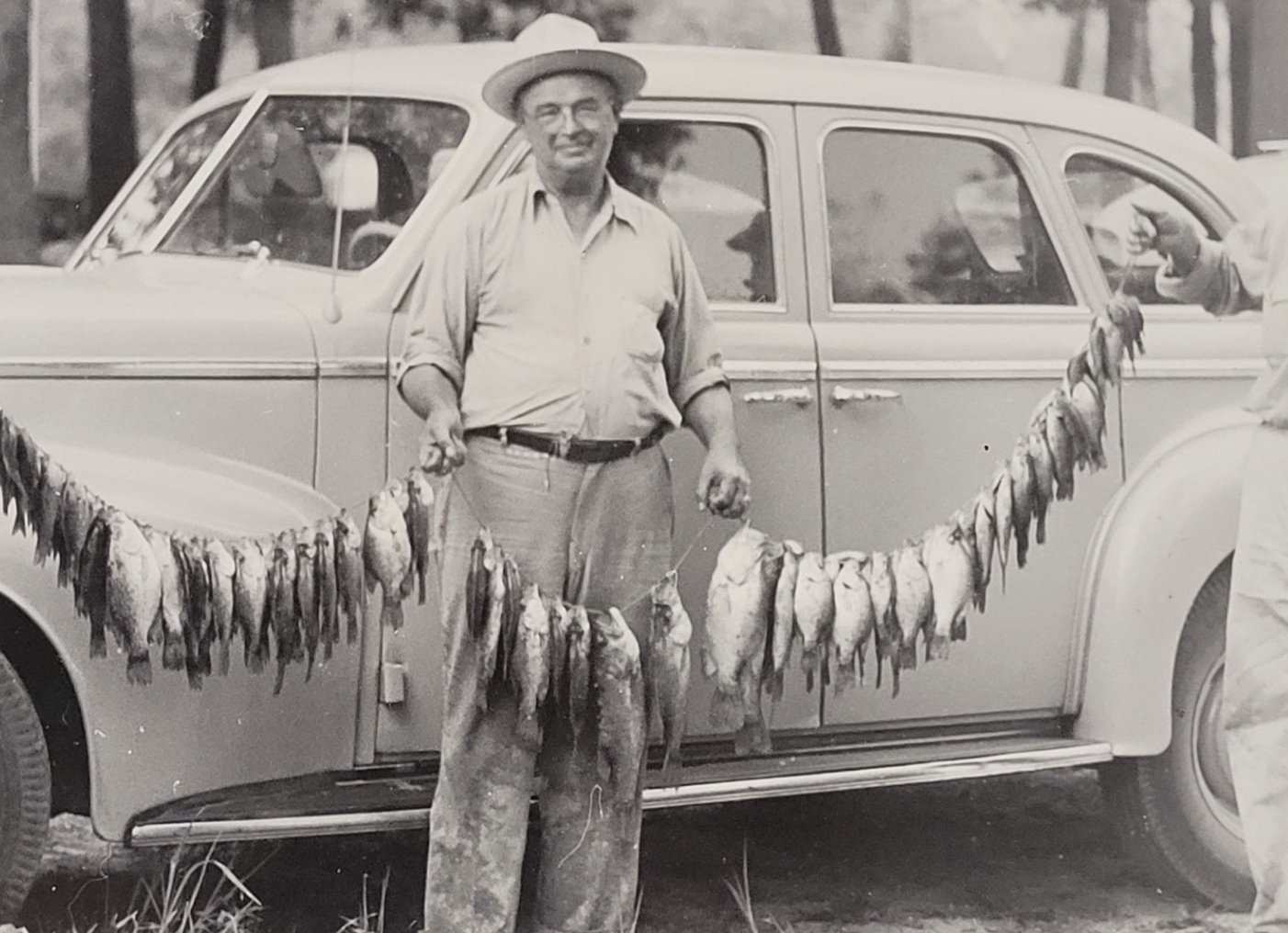 Historic black and white photo of man holding long stringer of fish. Wears light shirt and slacks with hat, standing in front of vintage car.