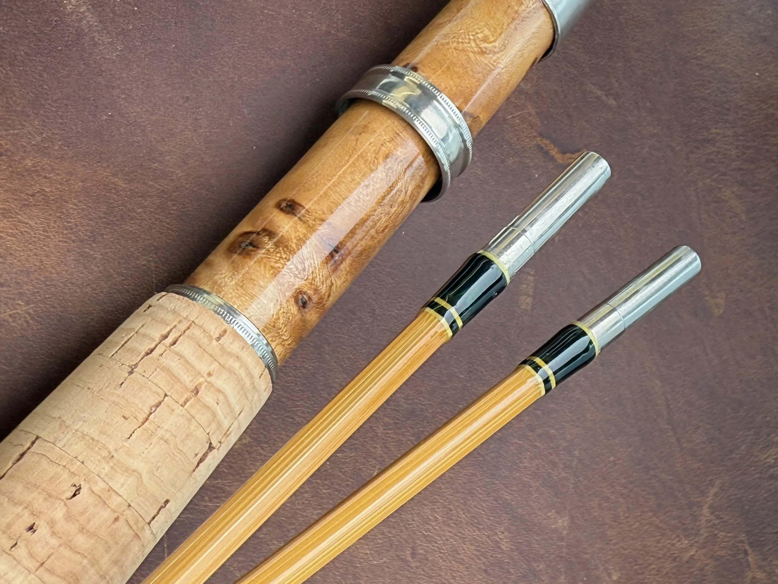Close-up of a handcrafted bamboo fly rod handle showing a highly figured honey-toned burl wood reel seat with a machined nickel silver slide band and a natural cork grip, alongside two light golden bamboo tip sections with dark green and cream thread