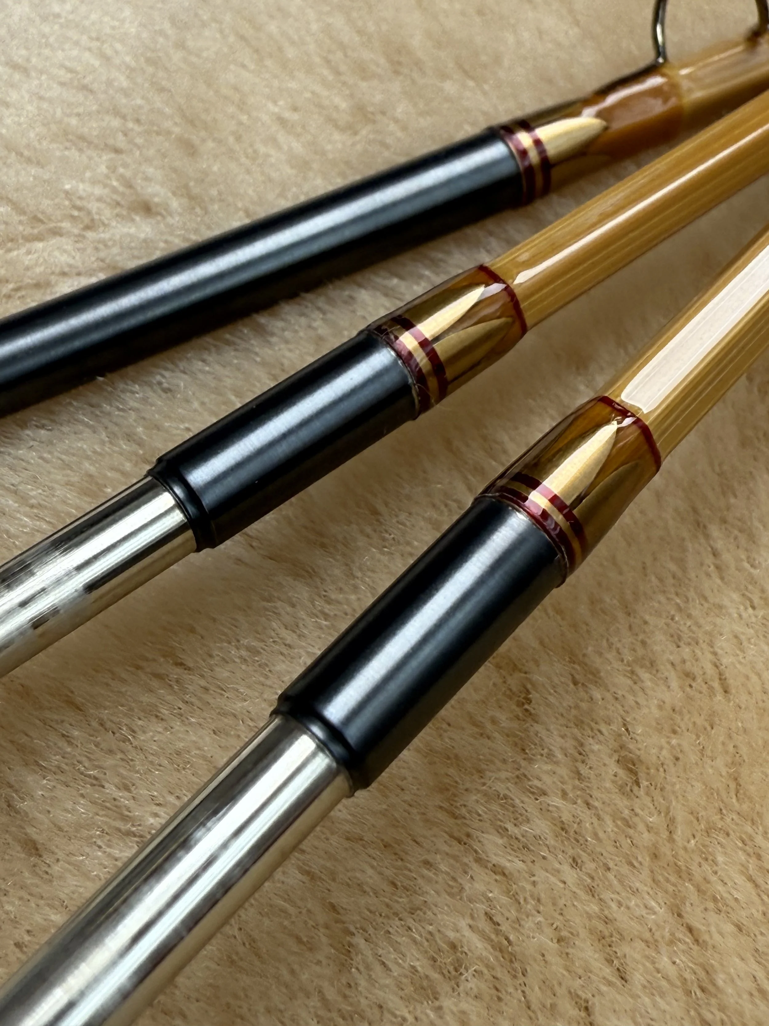 Macro close-up of three blued nickel silver bamboo fly rod ferrules, each with antique gold main wraps, red trim wraps and crowned tabs, displayed on shearling.