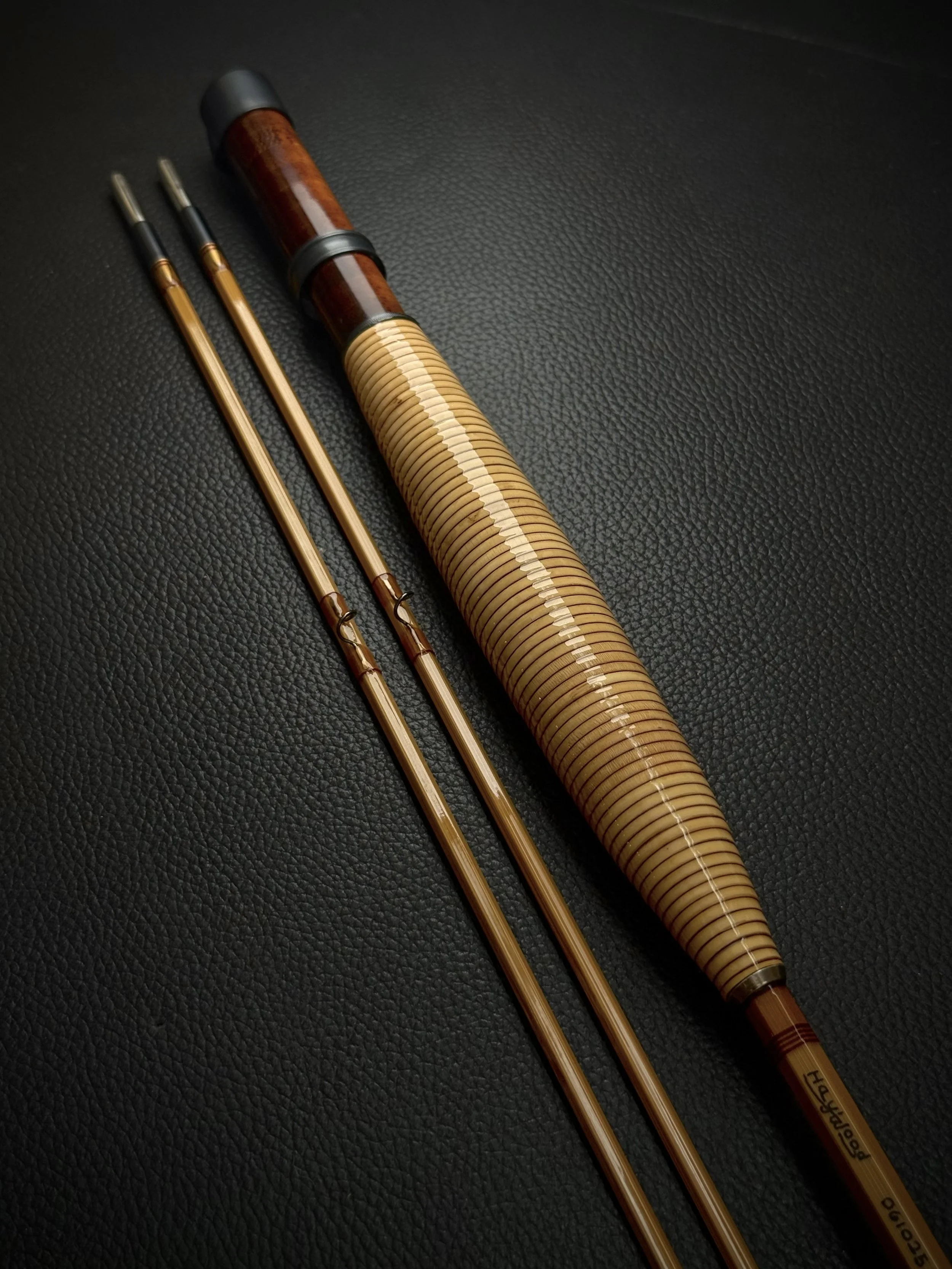 A Haywood bamboo fly rod with a rattan grip, a flamed bamboo reel seat with a blued hardware, and two slender blonde bamboo tip sections with small snake guides, displayed against a dark leather background.