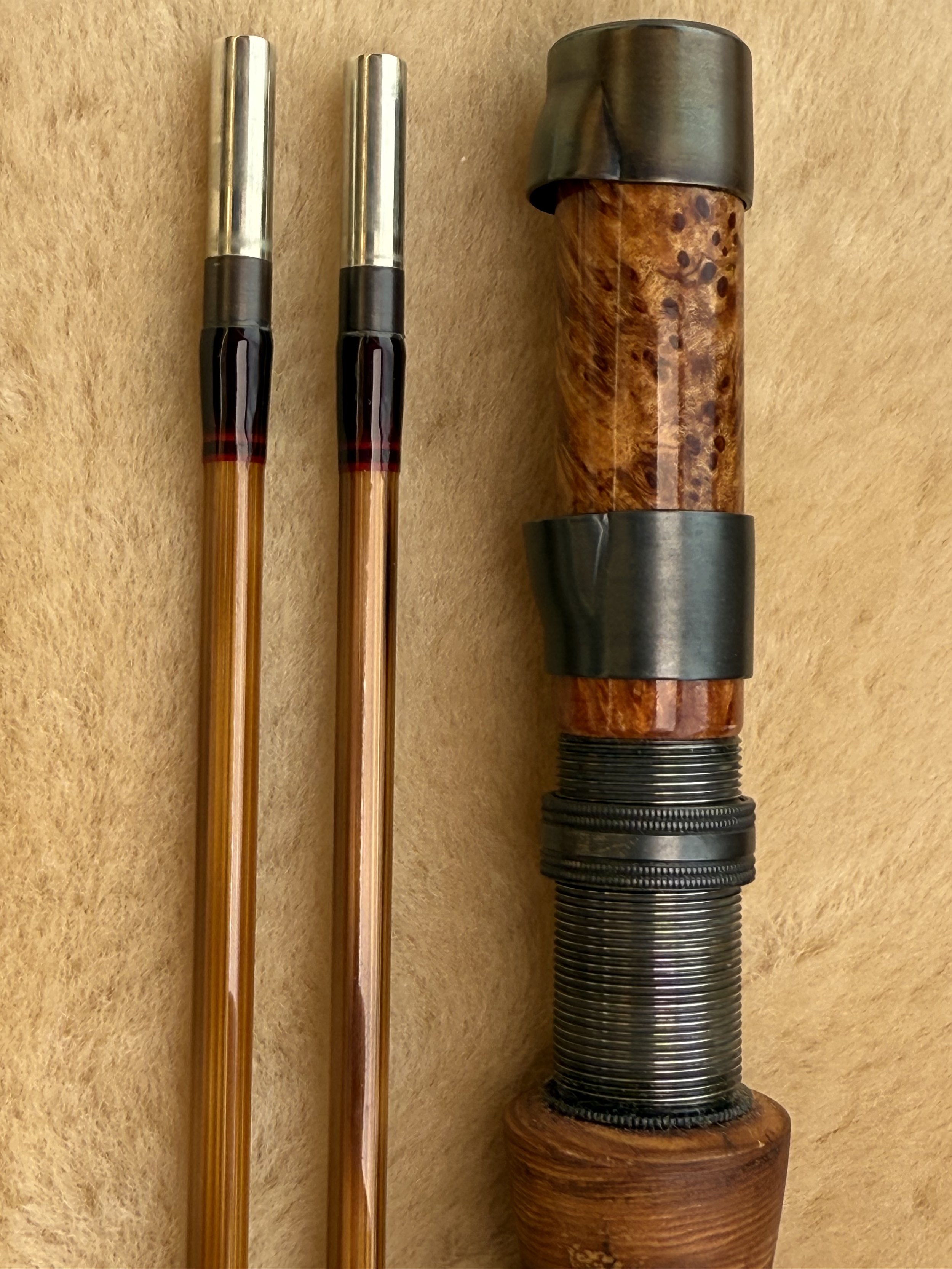 Close-up of a handcrafted fly rod reel seat featuring a highly figured burl wood insert flanked by blued hardware with a threaded locking collar, beside flamed cane rod sections with nickel silver ferrules and dark red thread trim wraps, on a sheepsk