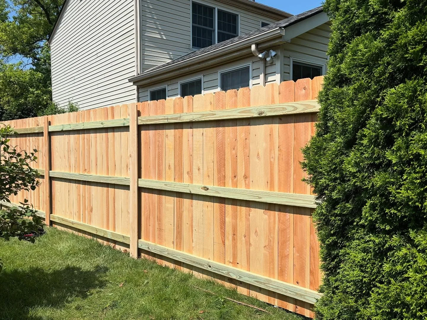 Backyard with a new wooden privacy fence, green grass, and a house with white siding and windows.