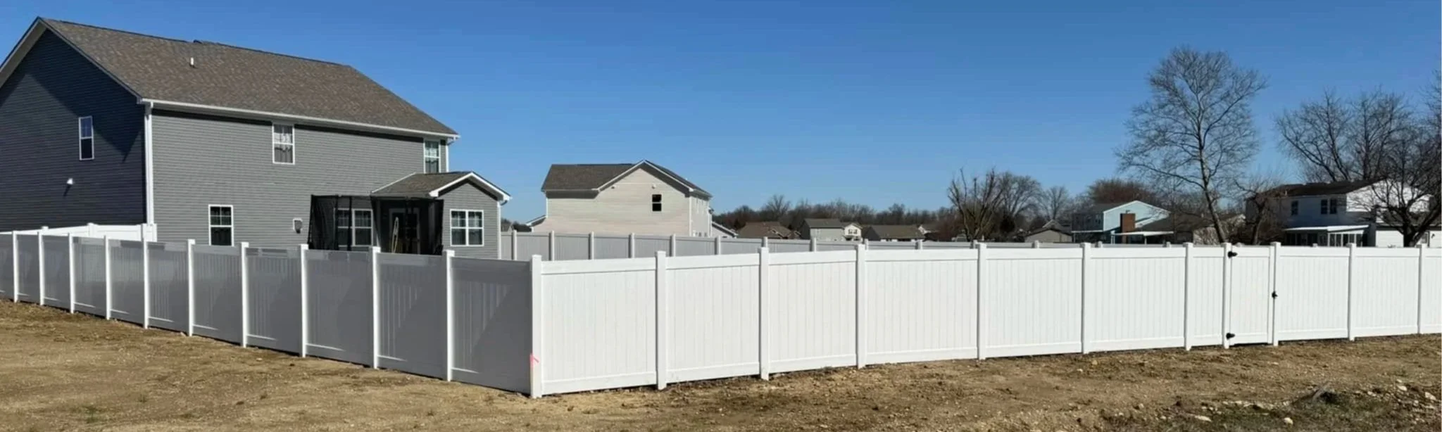 Photo of a backyard enclosed by a white vinyl privacy fence with houses and trees in the background under a clear blue sky.