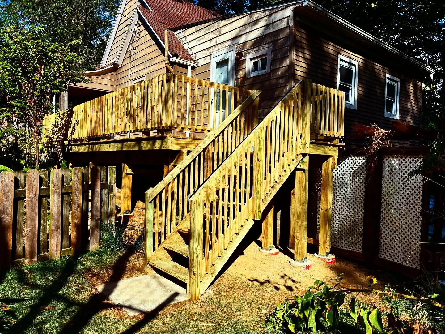 Wooden staircase leading up to the entrance of a brown house with a deck, surrounded by greenery and a wooden fence.
