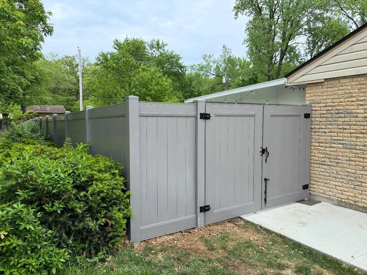 View of a gray privacy fence and gate next to a brick house, with green bushes and trees in the background.