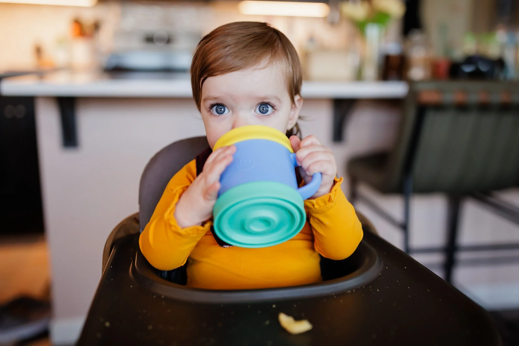 young toddler with blue eyes drinking out of colorful sippy cup in kitchen