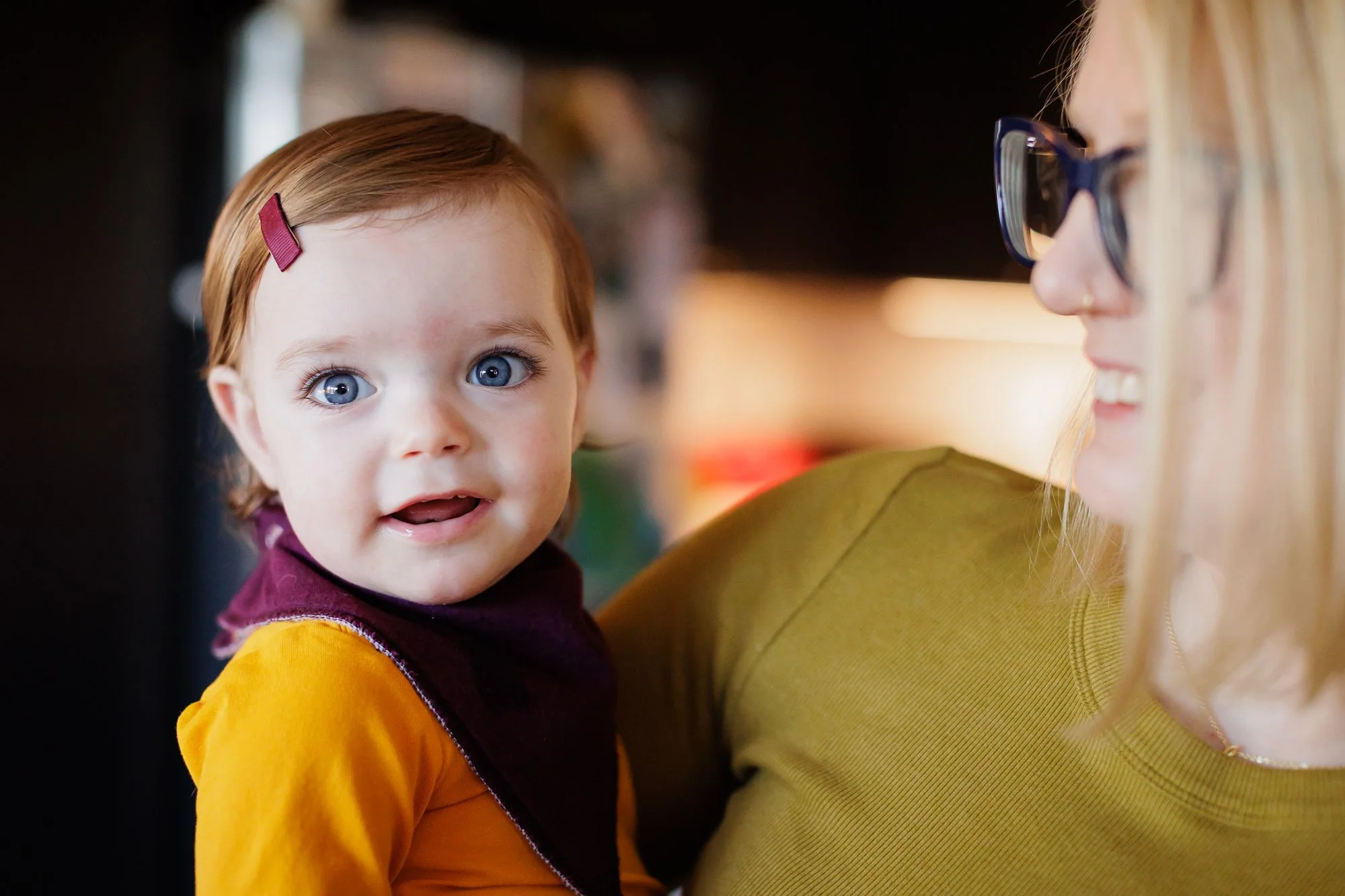 blue eyed toddler in yellow shirt smiles at photographer while held by mother