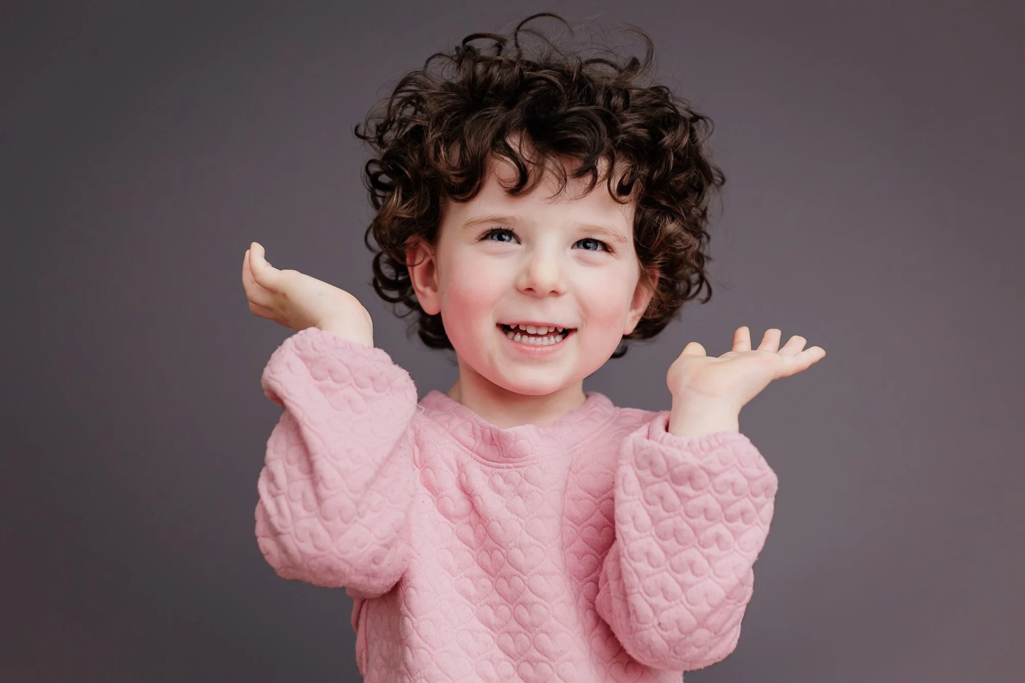 Toddler with brown curly hair and pink heart sweatshirt smiling for modern school photo