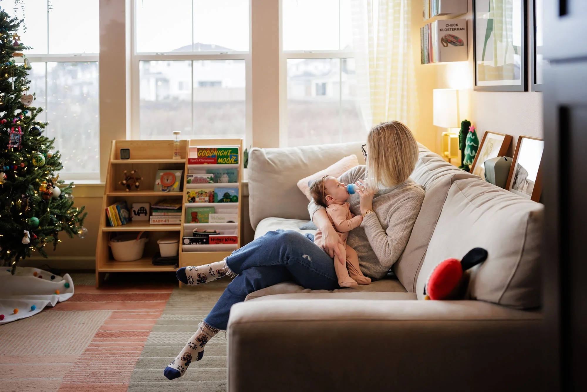 mom holding baby on couch feeding bottle with holiday tree in the background