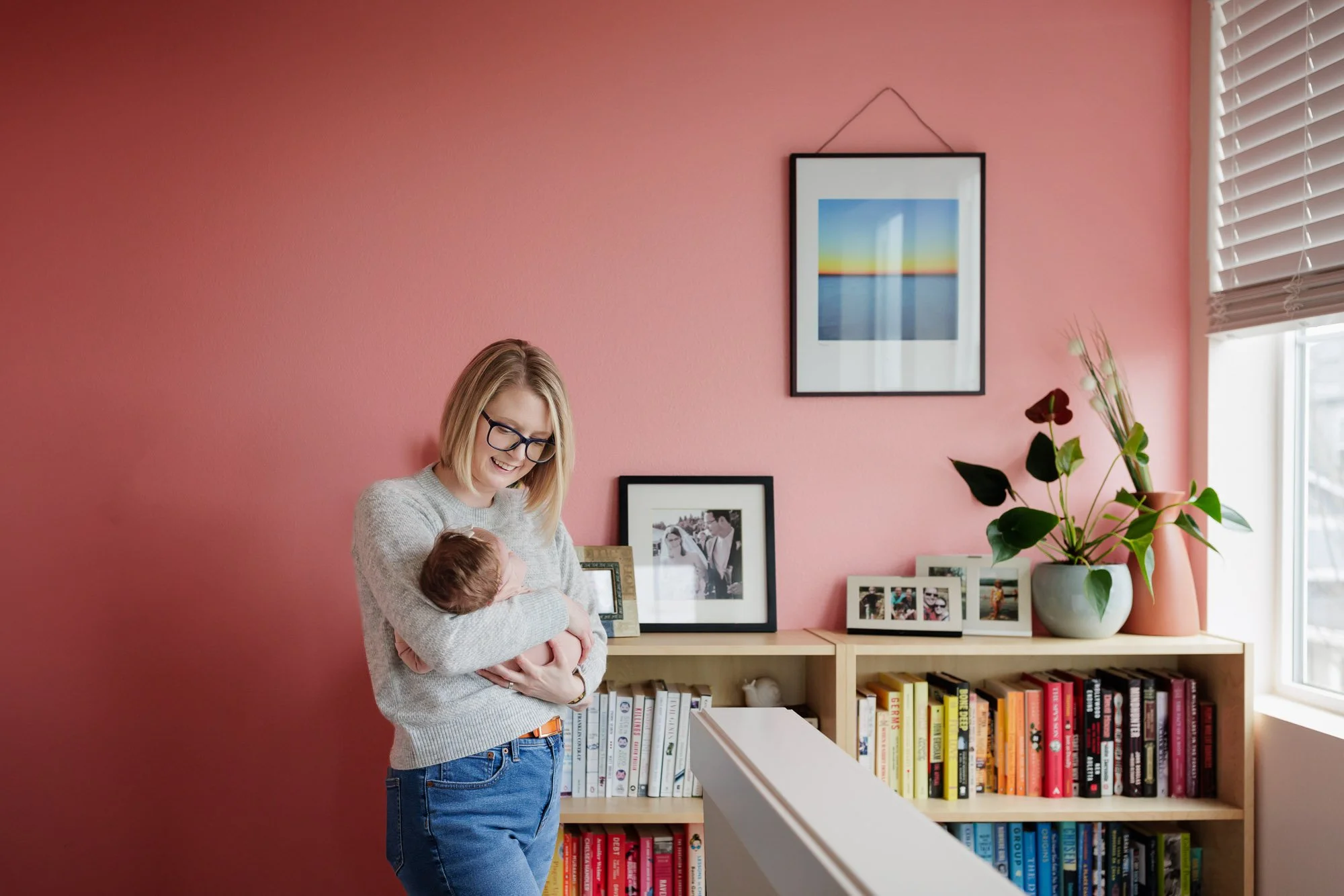 mom holds baby in pink colorful hallway by bookshelves and window