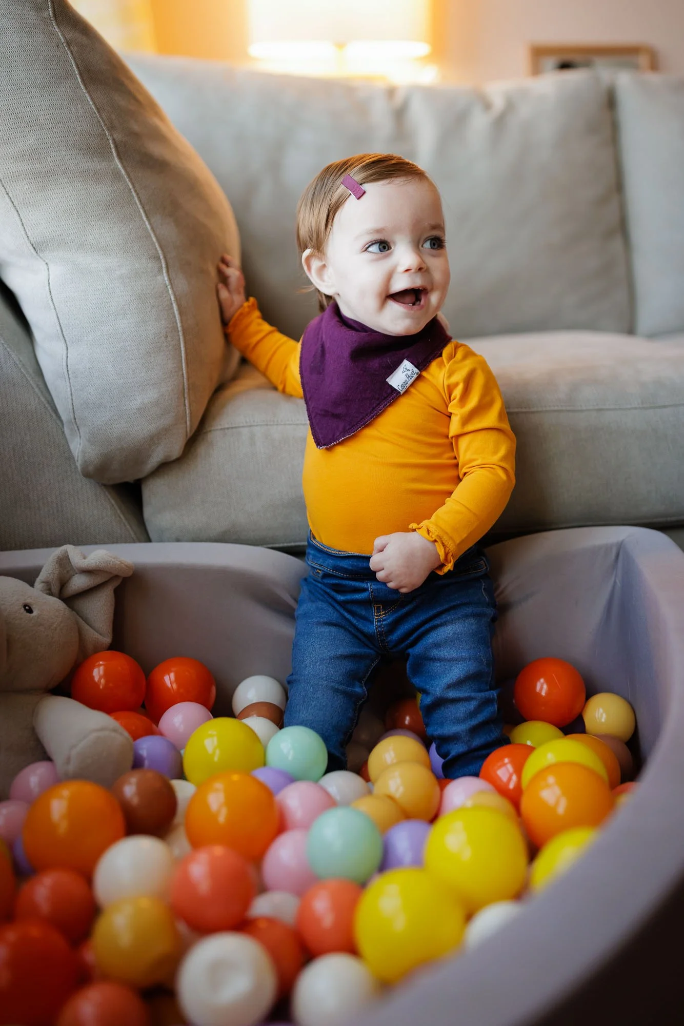 toddler in yellow shirt and jeans stands in colorful ball pit in living room