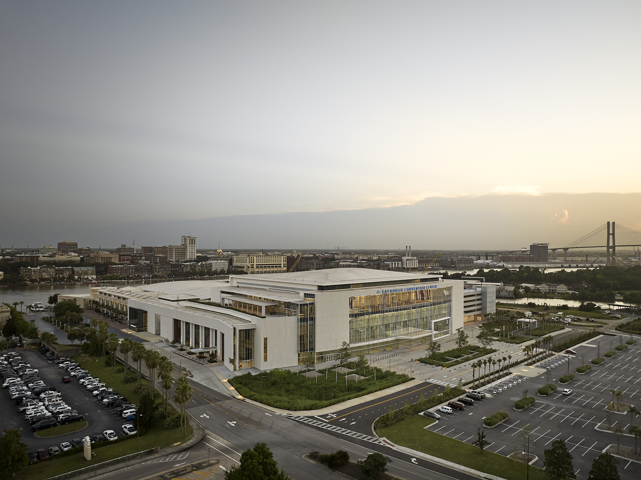 A wide-angle exterior architectural photograph of the Savannah Convention Center expansion at sunset, showing the glass facade overlooking the river and bridge
