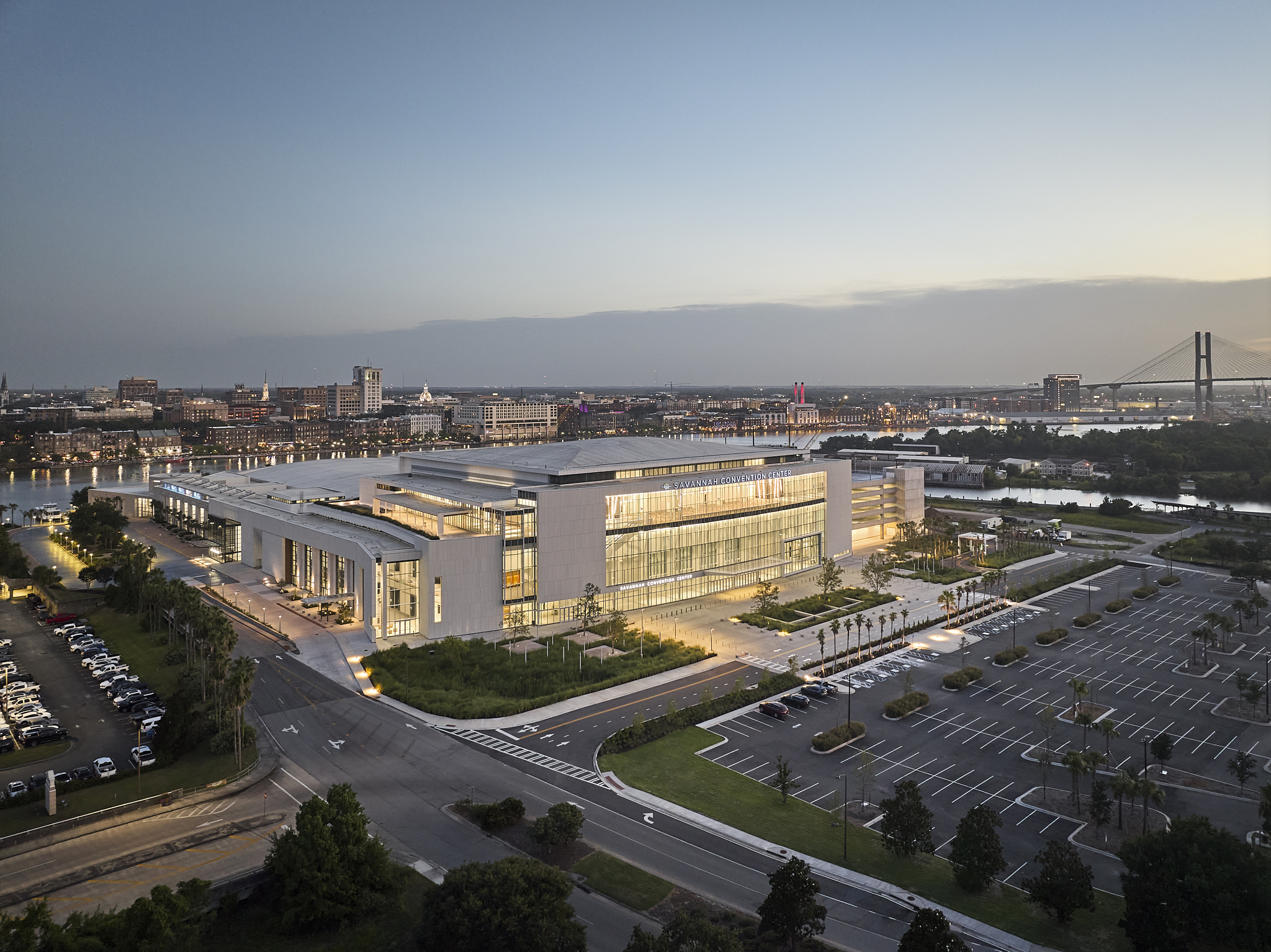 A wide-angle exterior architectural photograph of the Savannah Convention Center expansion at sunset, showing the glass facade and the Savannah Terrace overlooking the river.