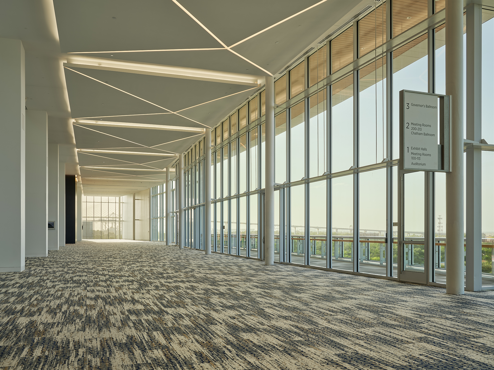 A wide-angle interior architectural photograph of the Savannah Convention Center expansion at sunset, showing the glass facade from the inside looking towards the bridge
