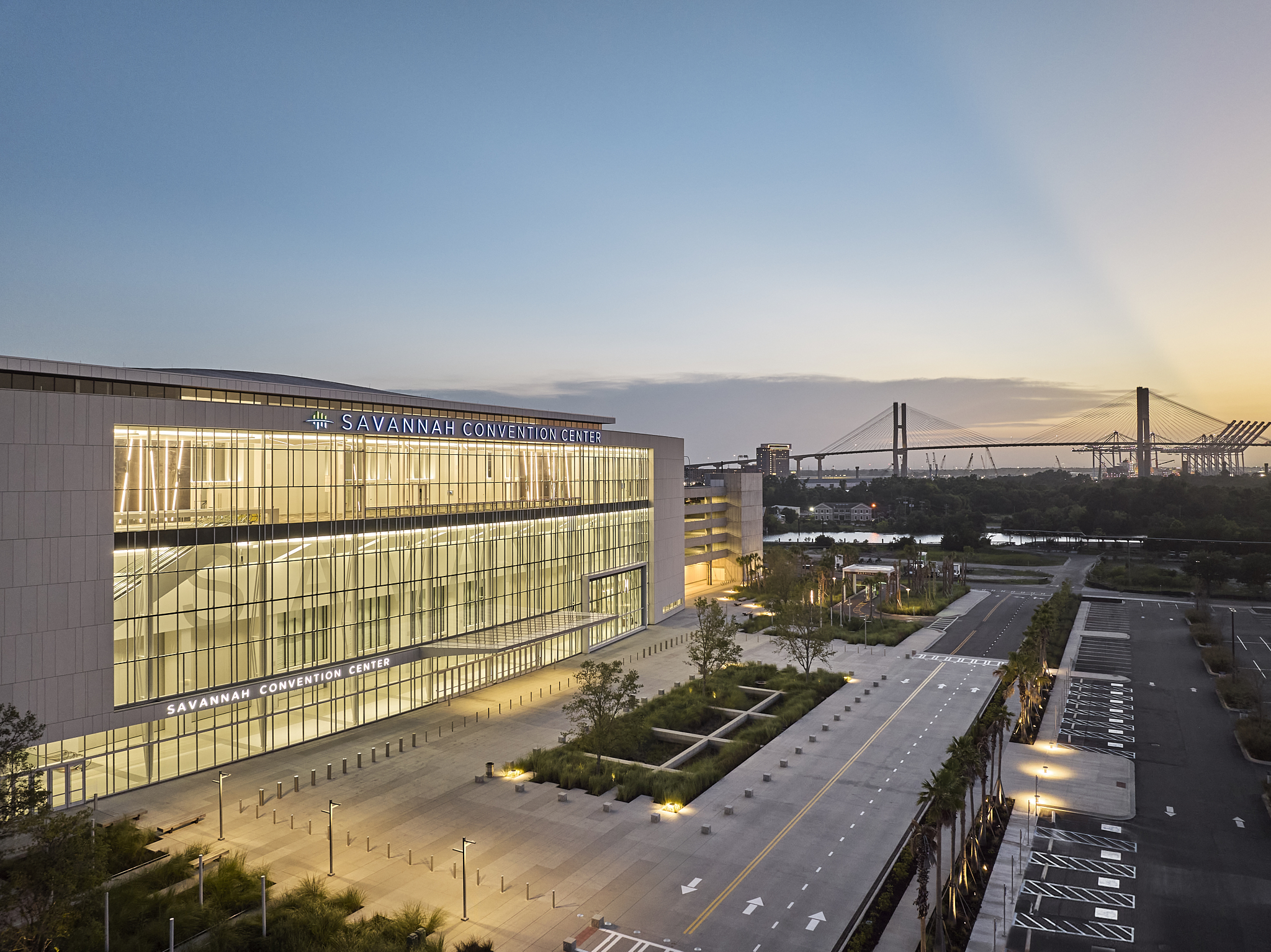A wide-angle exterior architectural photograph of the Savannah Convention Center expansion at sunset, showing the glass facade and the Savannah Terrace overlooking the bridge