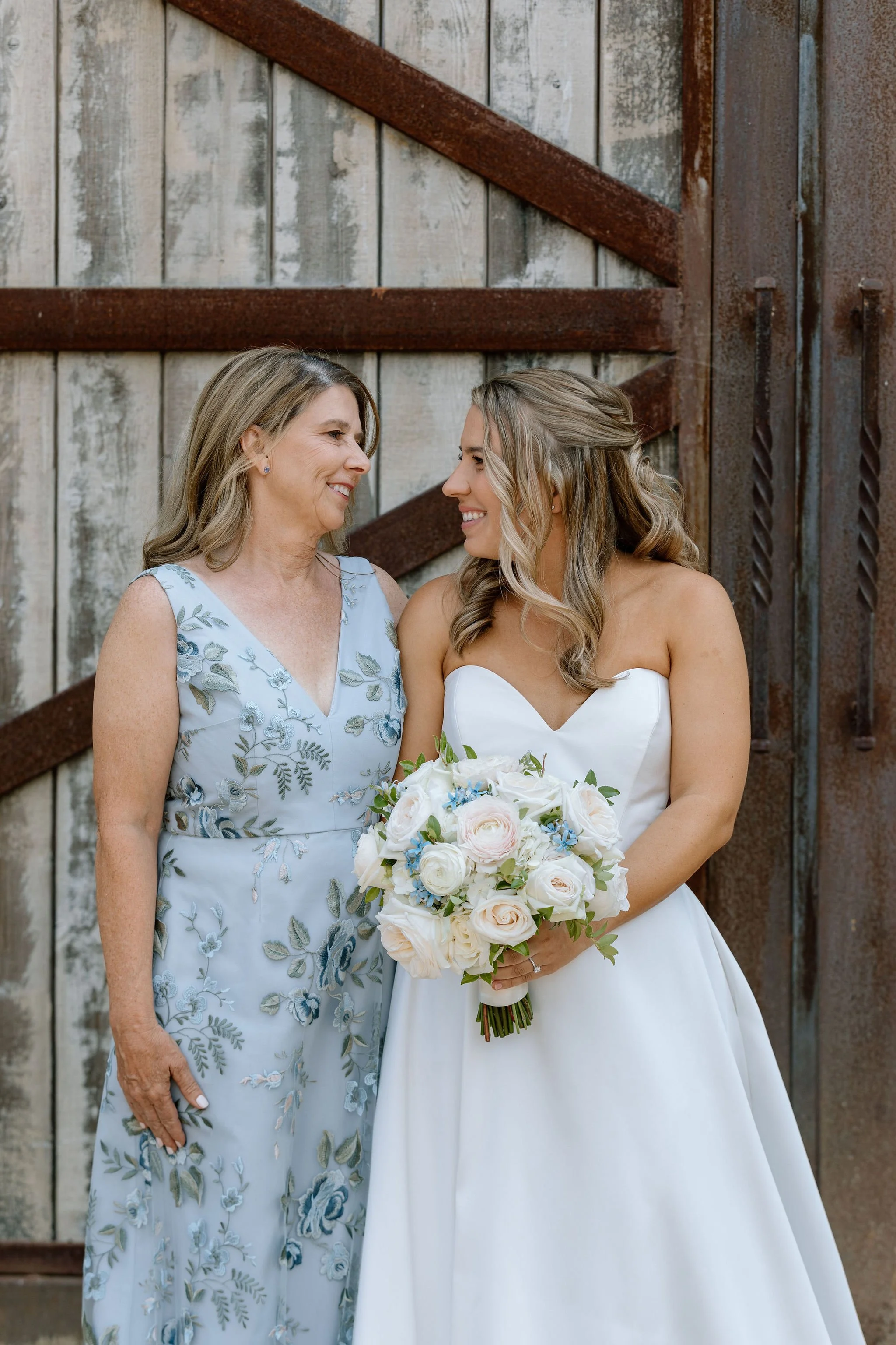 A smiling bride in a strapless white gown holds a lush bouquet of blush and white roses accented with soft blue blooms, standing beside her mother in an elegant floral dress against a rustic wooden backdrop in Sonoma County.