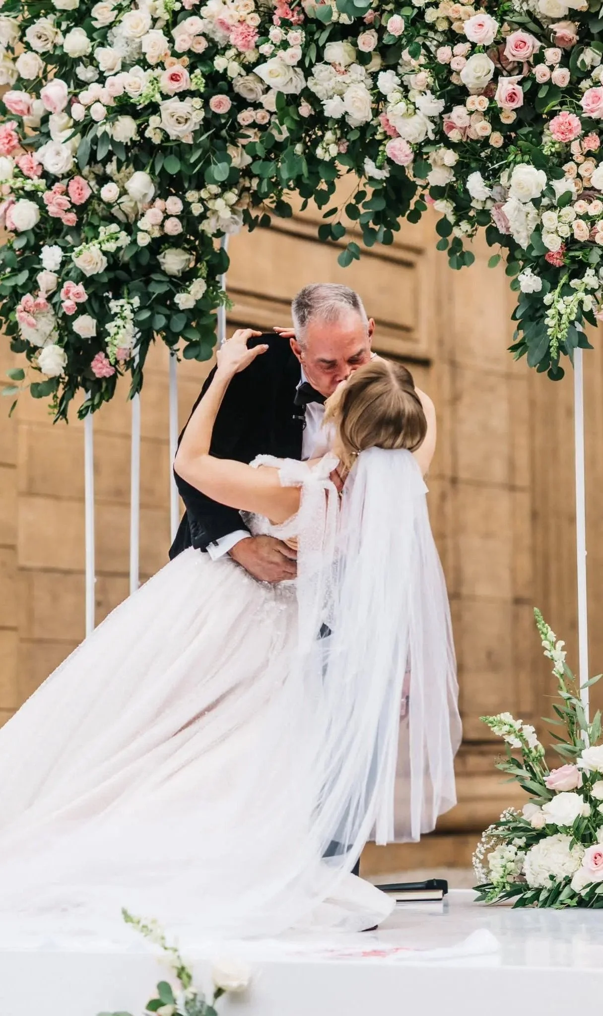 Bride and groom sharing a romantic kiss under a lush floral arch adorned with blush, pink, and white roses, accented with greenery, designed by a Santa Cruz-based florist.