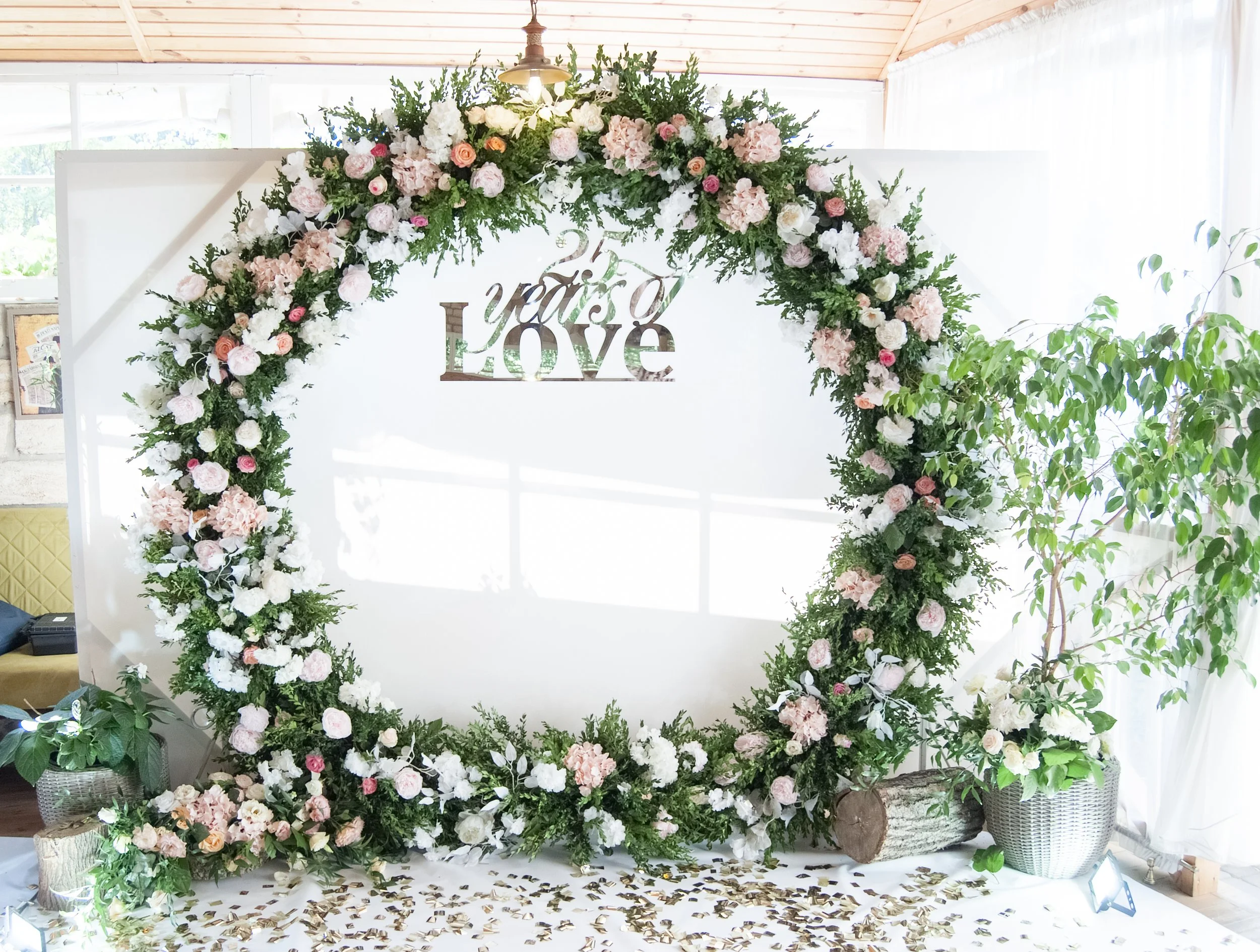 A round floral arch adorned with lush greenery, white, blush, and pink flowers, featuring a '25 years of love' sign in the center, complemented by potted plants and golden confetti on the floor.