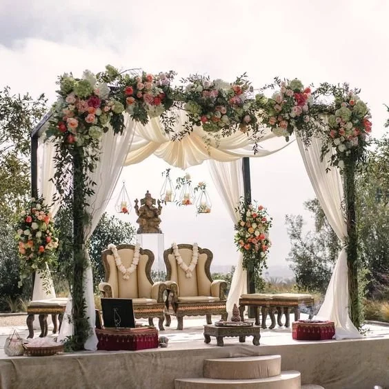 Luxurious wedding mandap decorated with pink, orange, and white flowers, cream drapes, and greenery. Elegant chairs and ceremonial items rest on a raised platform under the open sky.