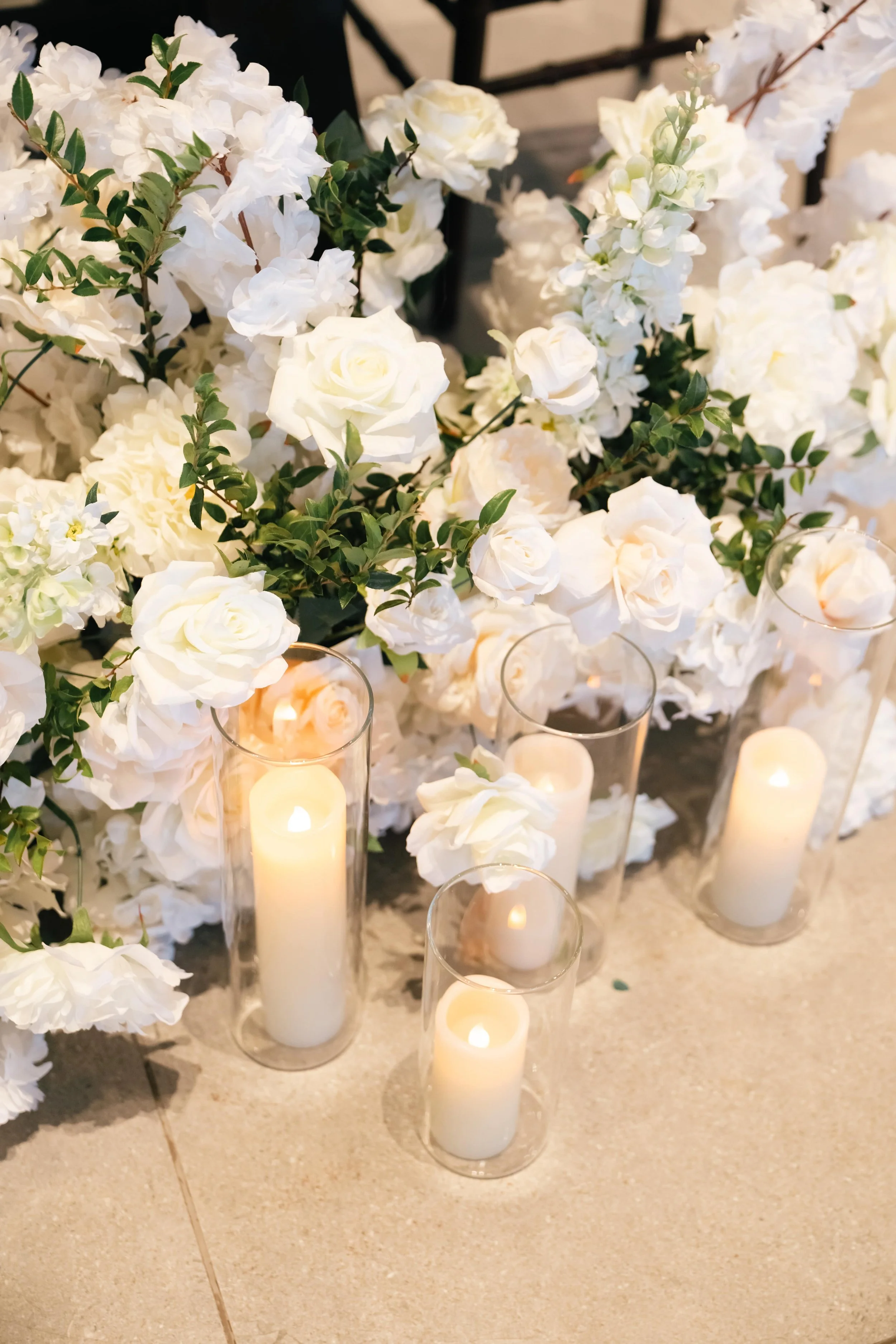 Elegant white floral arrangement featuring roses, lush greenery, and soft candlelight in cylindrical glass holders, creating a romantic wedding decor setting.