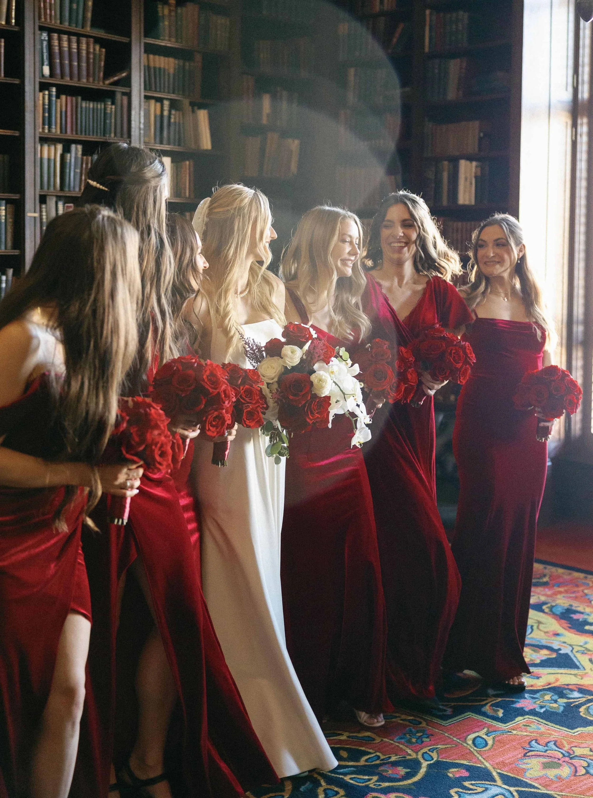 A joyful bride and her bridal party stand together, with the bride holding an elegant bouquet of red roses and white orchids, designed by a Napa County-based florist.