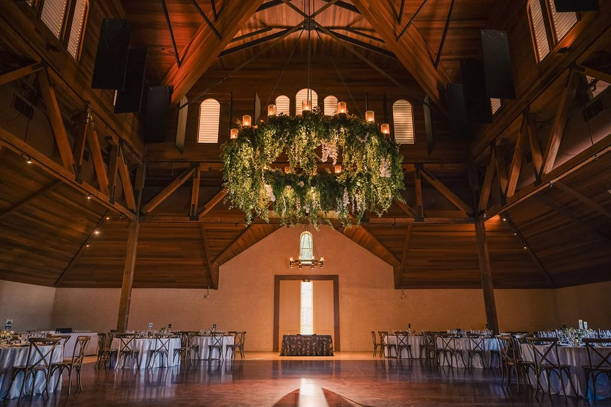 An elegant wedding reception hall with wooden beams and a large floral chandelier adorned with lush greenery and cascading white blooms, creating a romantic ambiance.