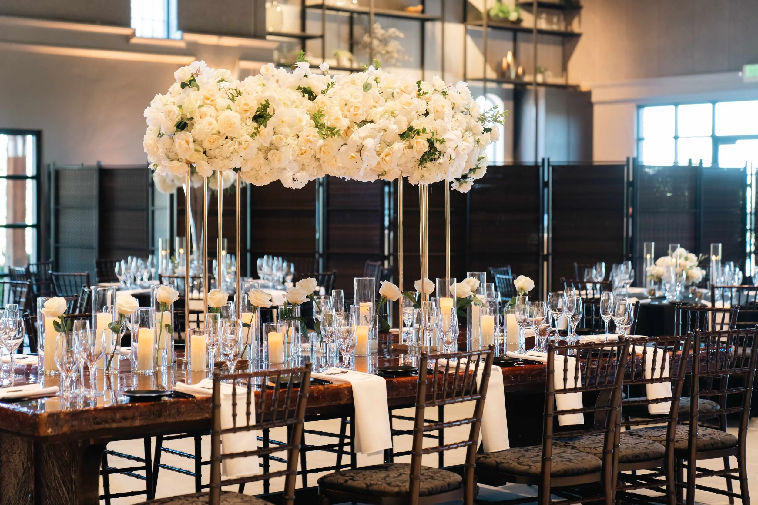 Elegant wedding reception setup featuring tall floral centerpieces of white roses and hydrangeas on gold stands, surrounded by glass candle holders and modern table settings on dark wooden tables.