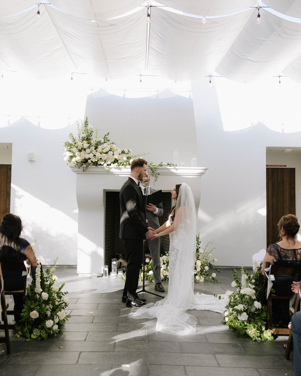 An elegant all-white palette transformed Blanco Urban Venue into a chic urban oasis for Bianca & John’s wedding. Lush roses, towering delphiniums, and touches of soft greenery lined the aisle, creating a bright, modern pathway toward the fireplace al