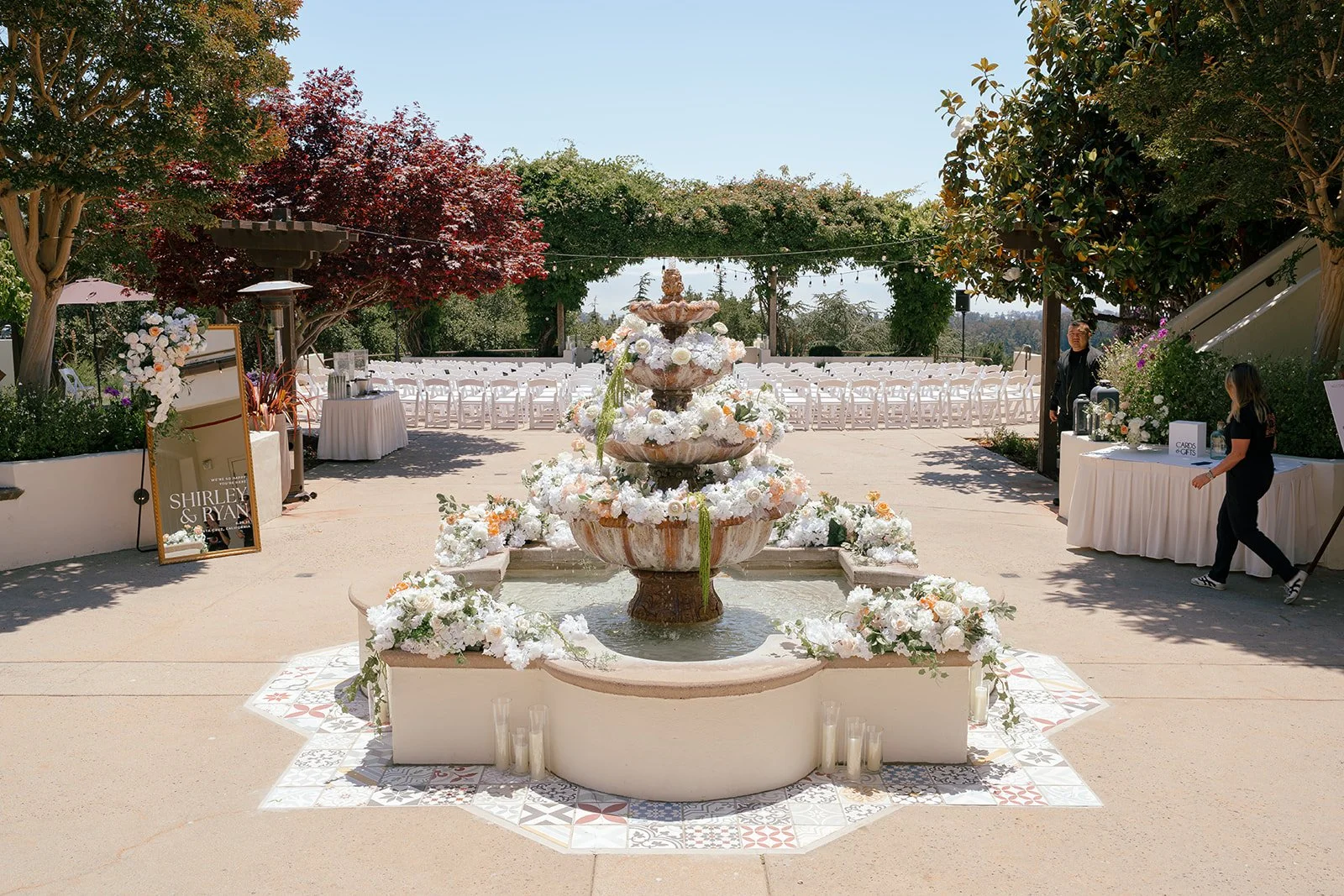 Outdoor wedding ceremony setup with a central fountain decorated with white and peach flowers, surrounded by white chairs, floral arrangements, and reception tables under trees on a sunny day.