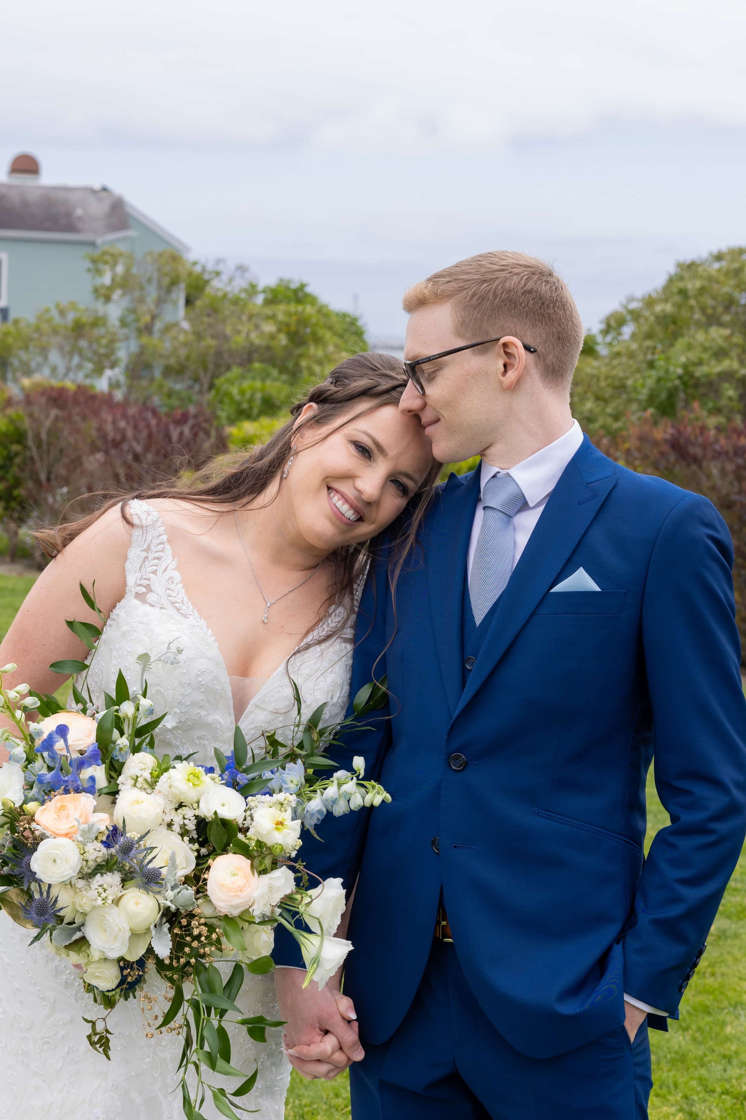 A bride holding a lush white and blue bouquet leans affectionately against her groom in a tailored navy blue suit, sharing a joyful moment in a serene garden setting in Napa County.