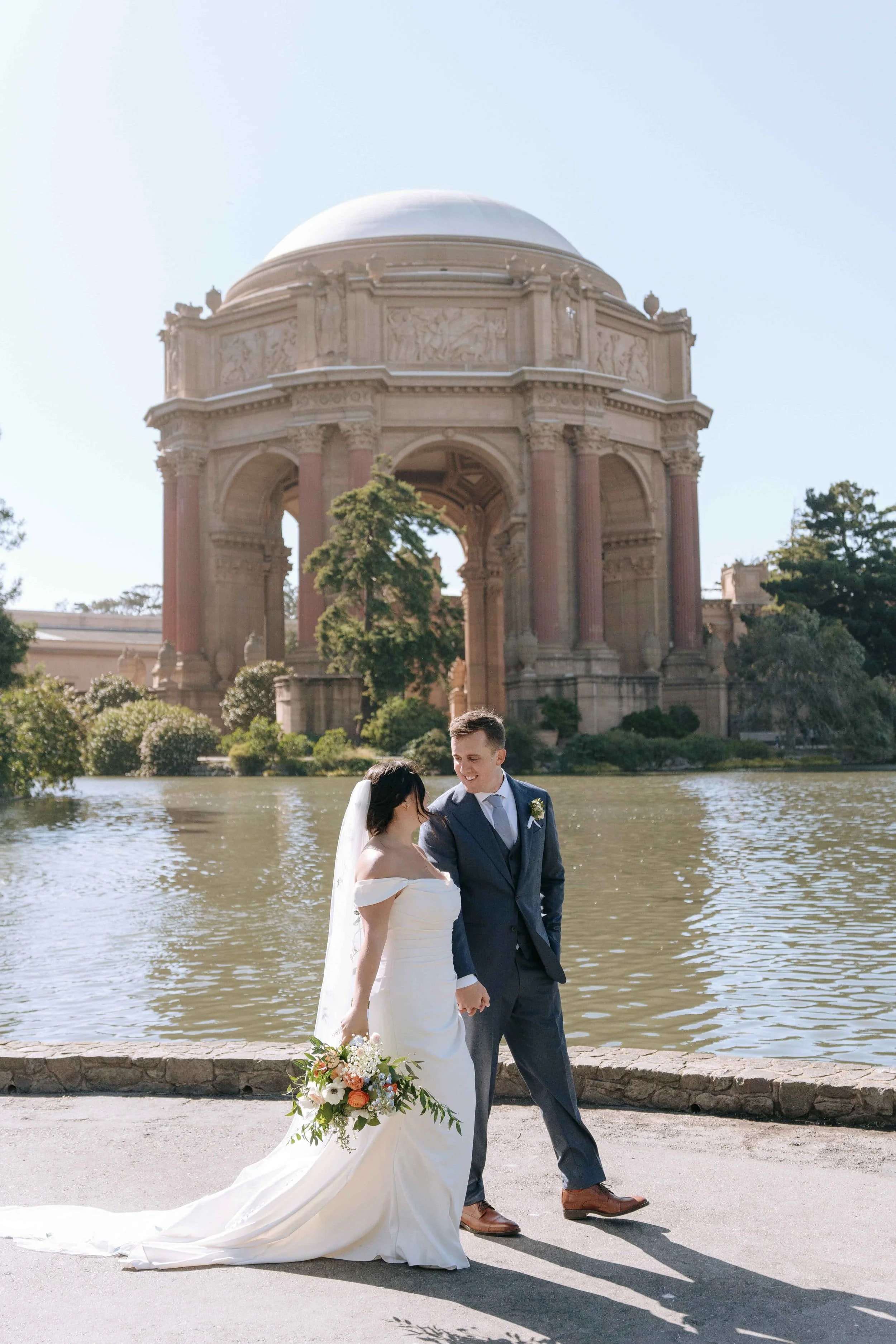 A bride and groom holding hands and smiling at each other in front of a large historic monument with a water body in front.