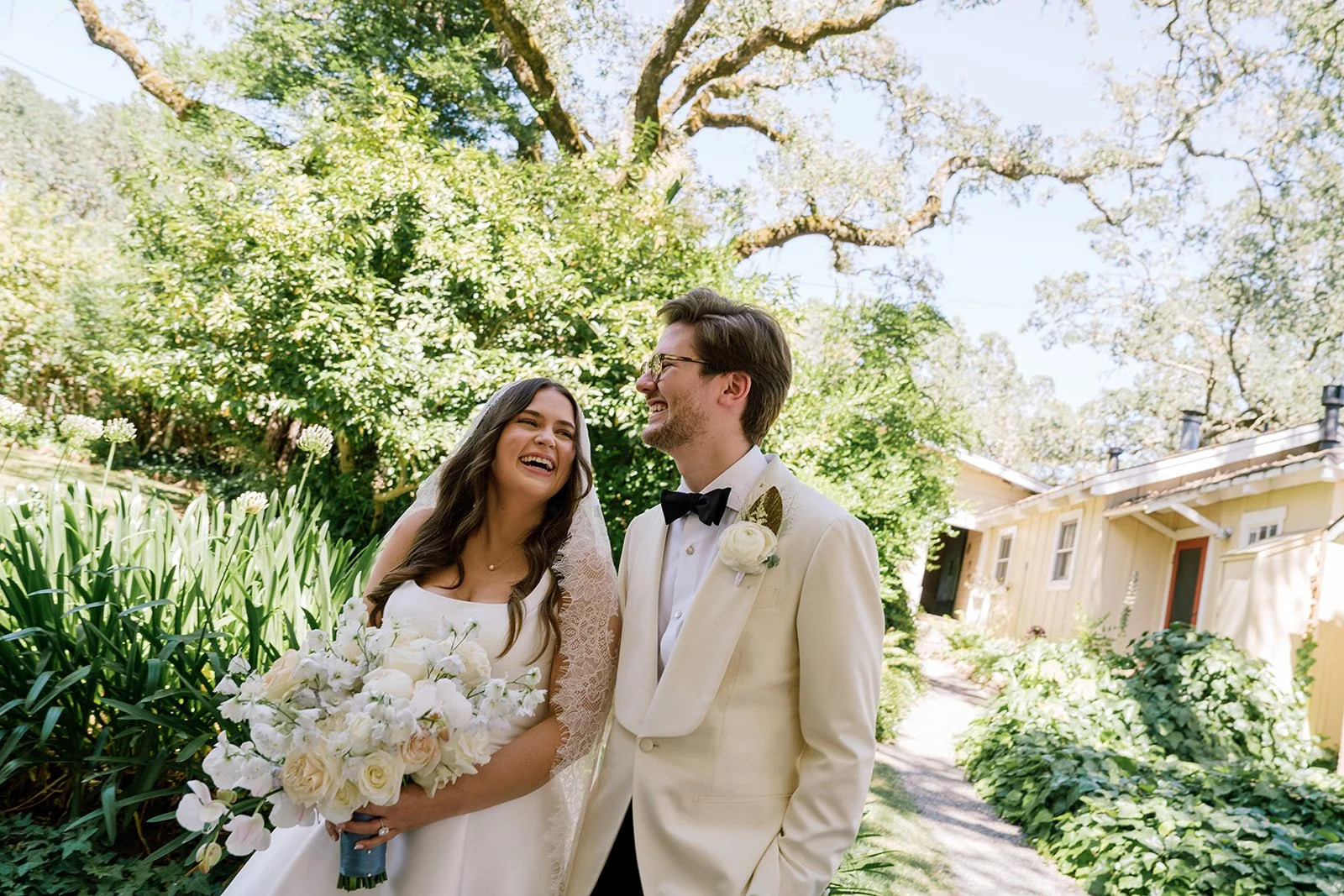 A bride and groom laughing outdoors in a garden setting on their wedding day, with greenery and a yellow house in the background.