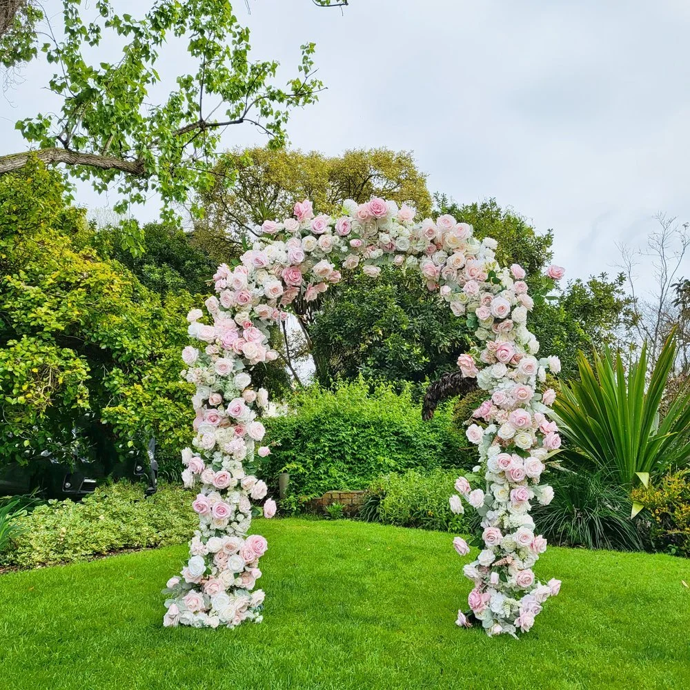 Semicircular floral arch beautifully adorned with pink and white roses, set on a lush green lawn with vibrant trees and foliage in the background, creating a picturesque outdoor wedding backdrop.