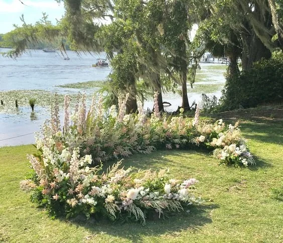 Ground floral arch design featuring soft pink and white blooms mixed with lush greenery, arranged in a semi-circle on a grassy lakeside setting, with trees draped in Spanish moss and serene water views in the background.