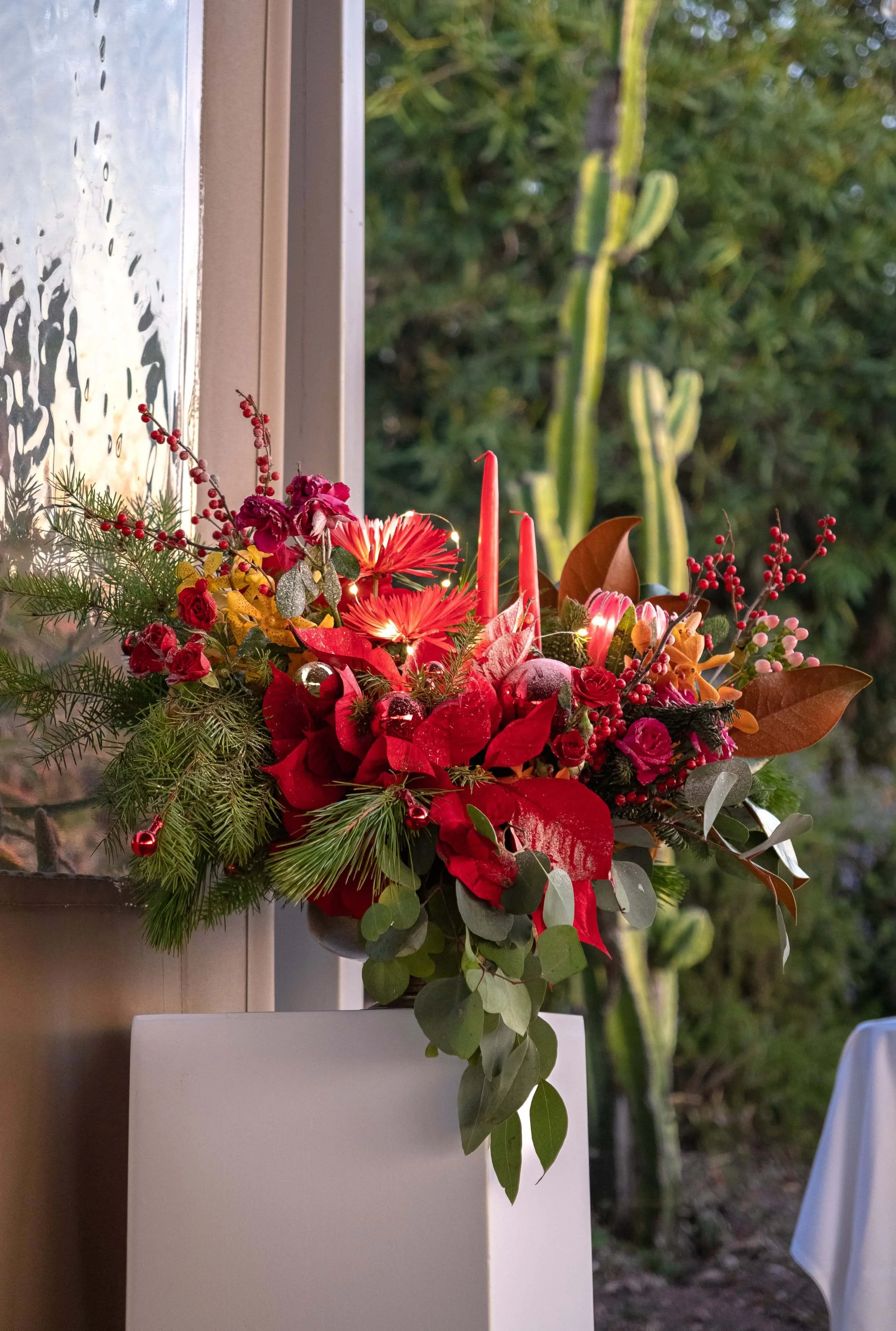 Festive floral arrangement featuring vibrant red poinsettias, candles, winter greenery, and seasonal accents, placed on a white pedestal with a natural outdoor background.