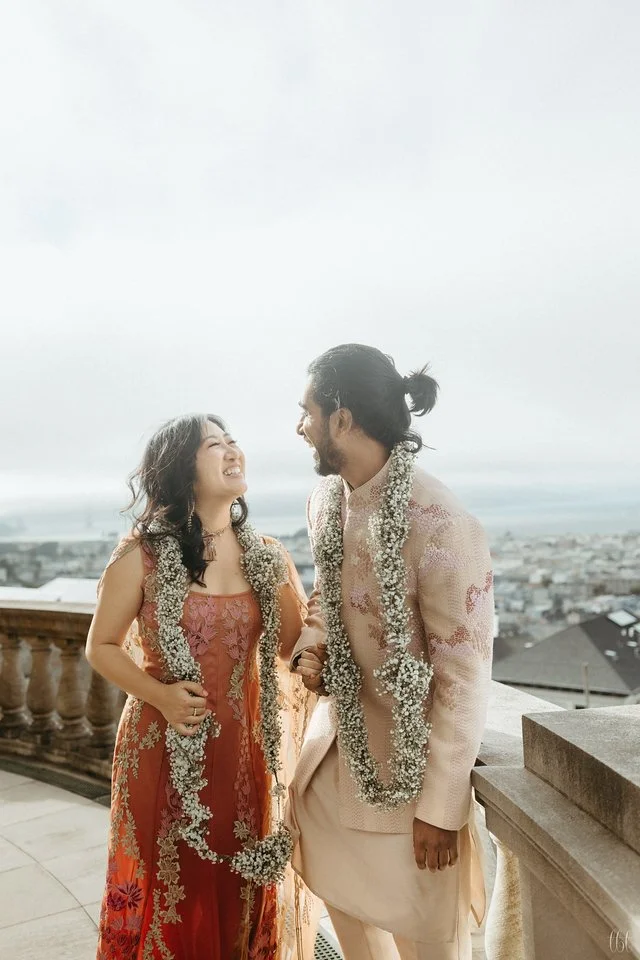 A joyful couple adorned in traditional floral garlands, designed by a San Francisco-based florist, share a heartfelt moment on a sunlit terrace.