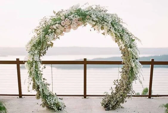 Moongate floral arch elegantly decorated with cascading greenery, white blooms, and blush roses, set against a serene waterfront backdrop.