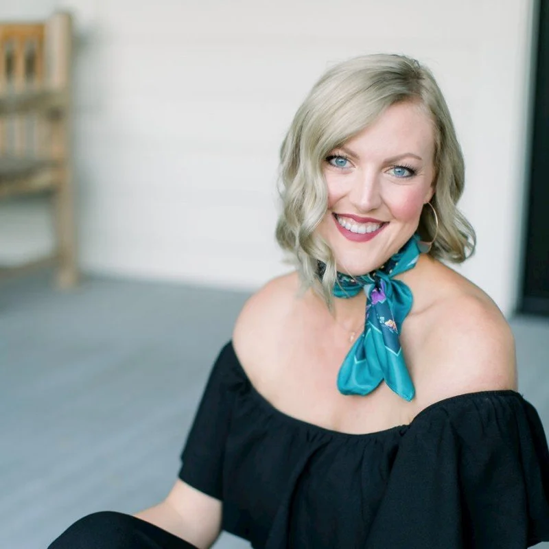 A smiling woman with shoulder-length blonde hair, blue eyes, wearing a black off-shoulder top and a teal silk scarf tied around her neck, sitting indoors with a blurred background.