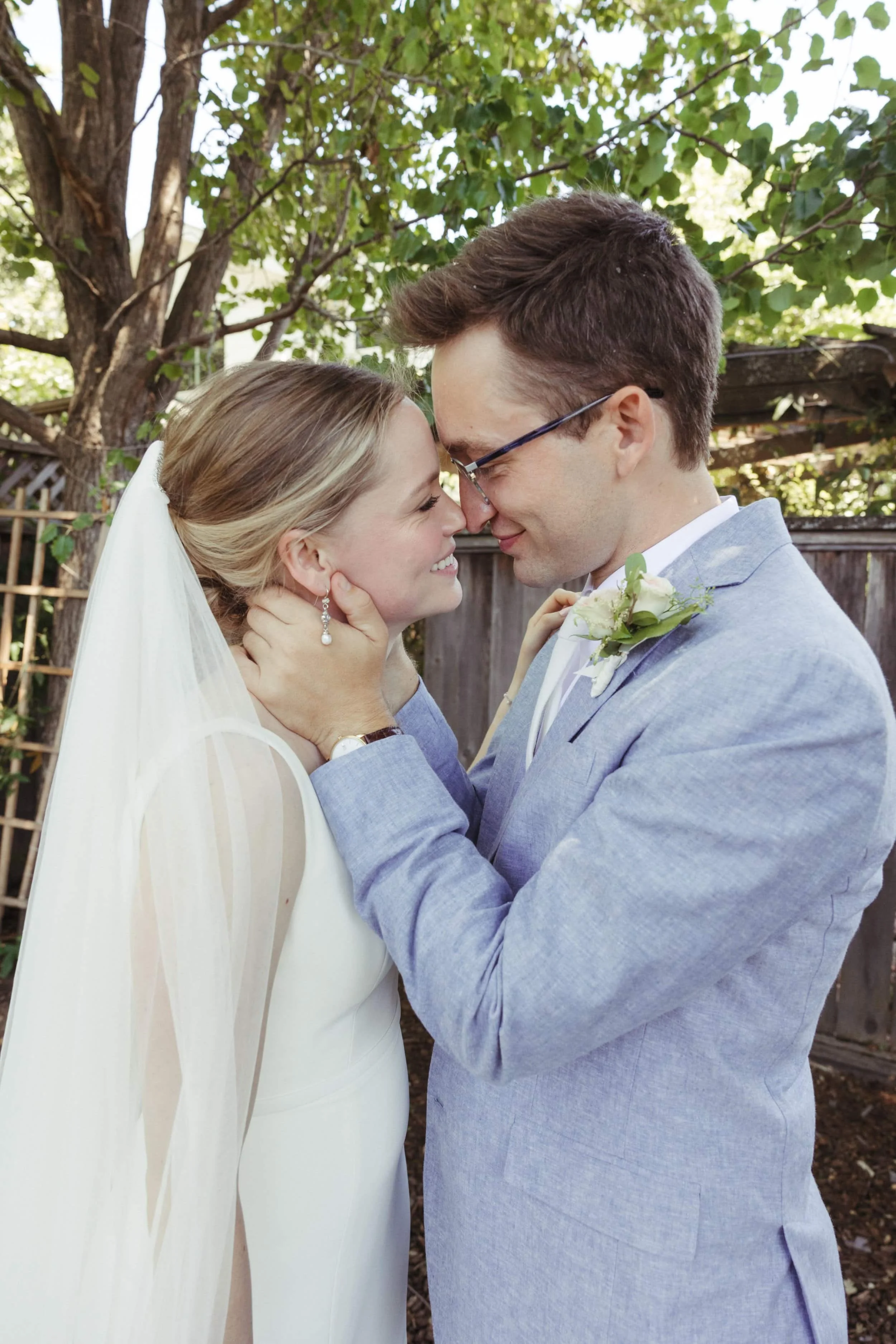 A joyful bride in a white gown and veil shares a tender moment with the groom, dressed in a light blue suit with a white boutonniere, as they smile and embrace beneath a canopy of trees in Sonoma County.