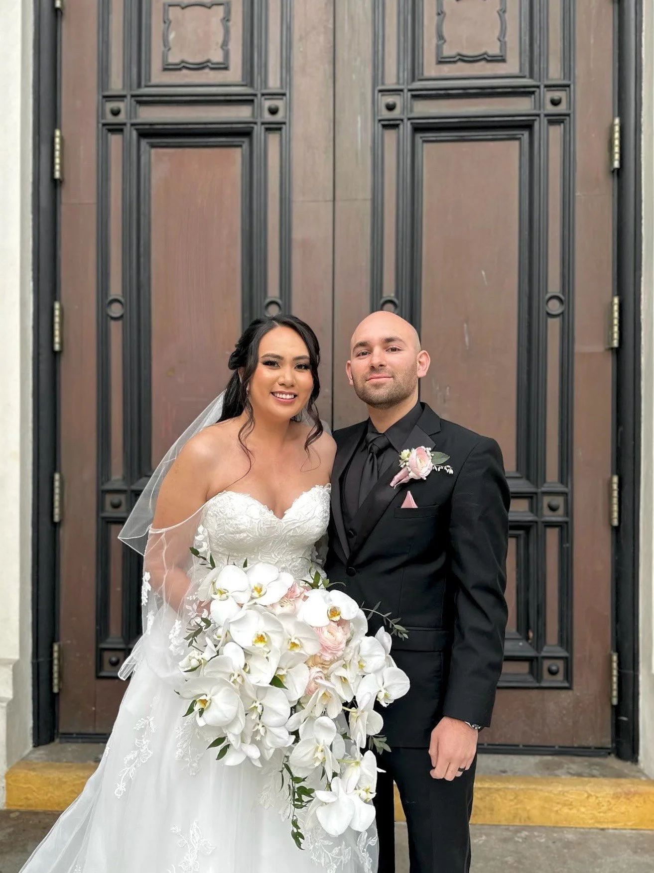 A joyful bride holding a stunning white orchid bouquet, designed by a Santa Rosa-based florist, poses with her groom in front of elegant wooden doors, both beaming with happiness.