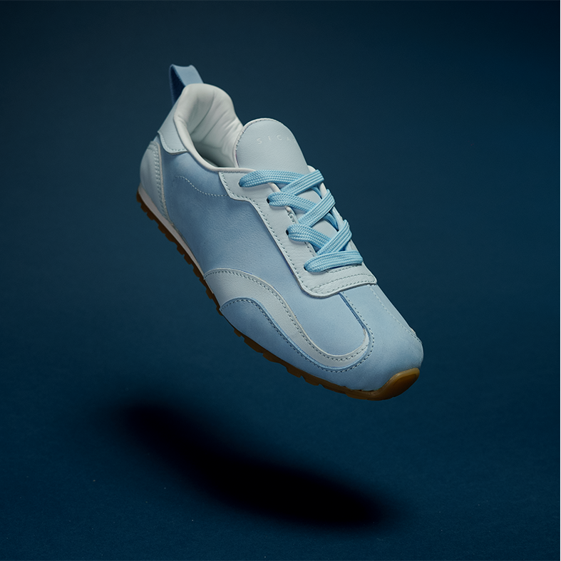 RINA Sneakerina Cielo colorway — sky blue sanded suede upper with gum sole and minimal foil stamp branding, floating against a dark studio background
