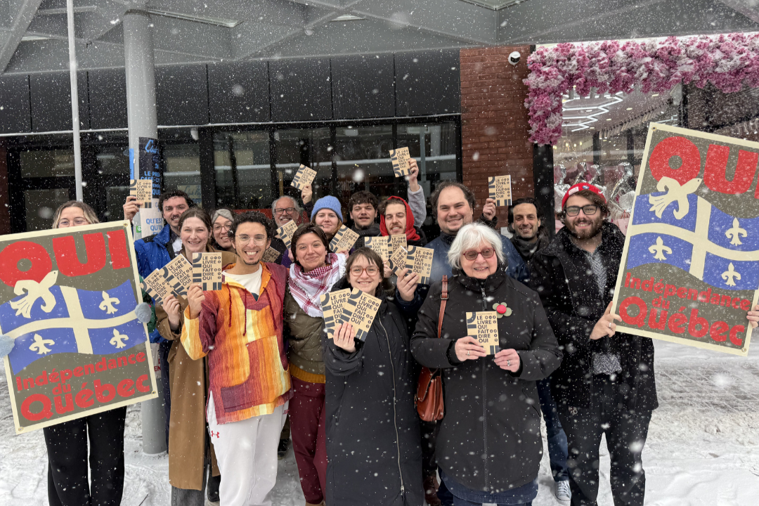 Groupe de personnes tenant des affiches et des flyers, célébrant la fête nationale du Québec, sous la neige, avec des décorations florales et un bâtiment moderne en arrière-plan.