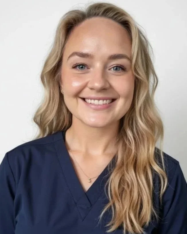 A young woman with long wavy blonde hair, blue eyes, and light skin, smiling at the camera, wearing dark blue medical scrubs.