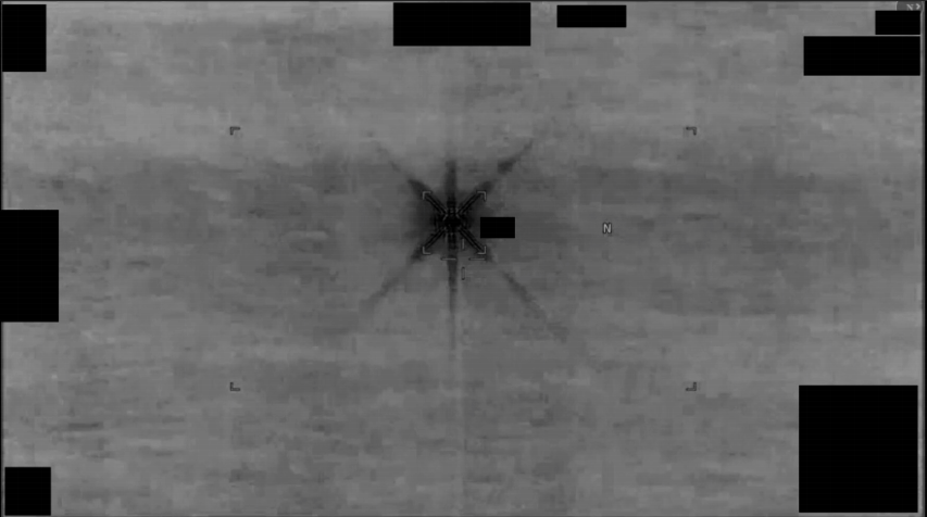 Grayscale infrared sensor footage from a U.S. military platform, showing a stationary target reticle centered on a small dark object against a mottled gray thermal background, with corner redaction blocks obscuring platform-identifying HUD data.
