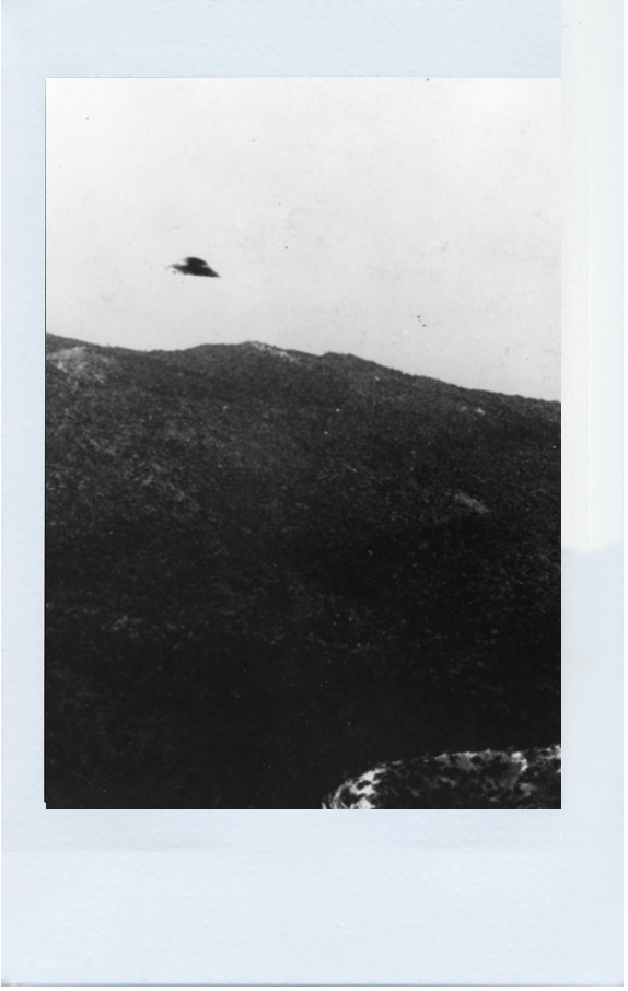Black and white image of a UFO flying in the sky above a hilly landscape.